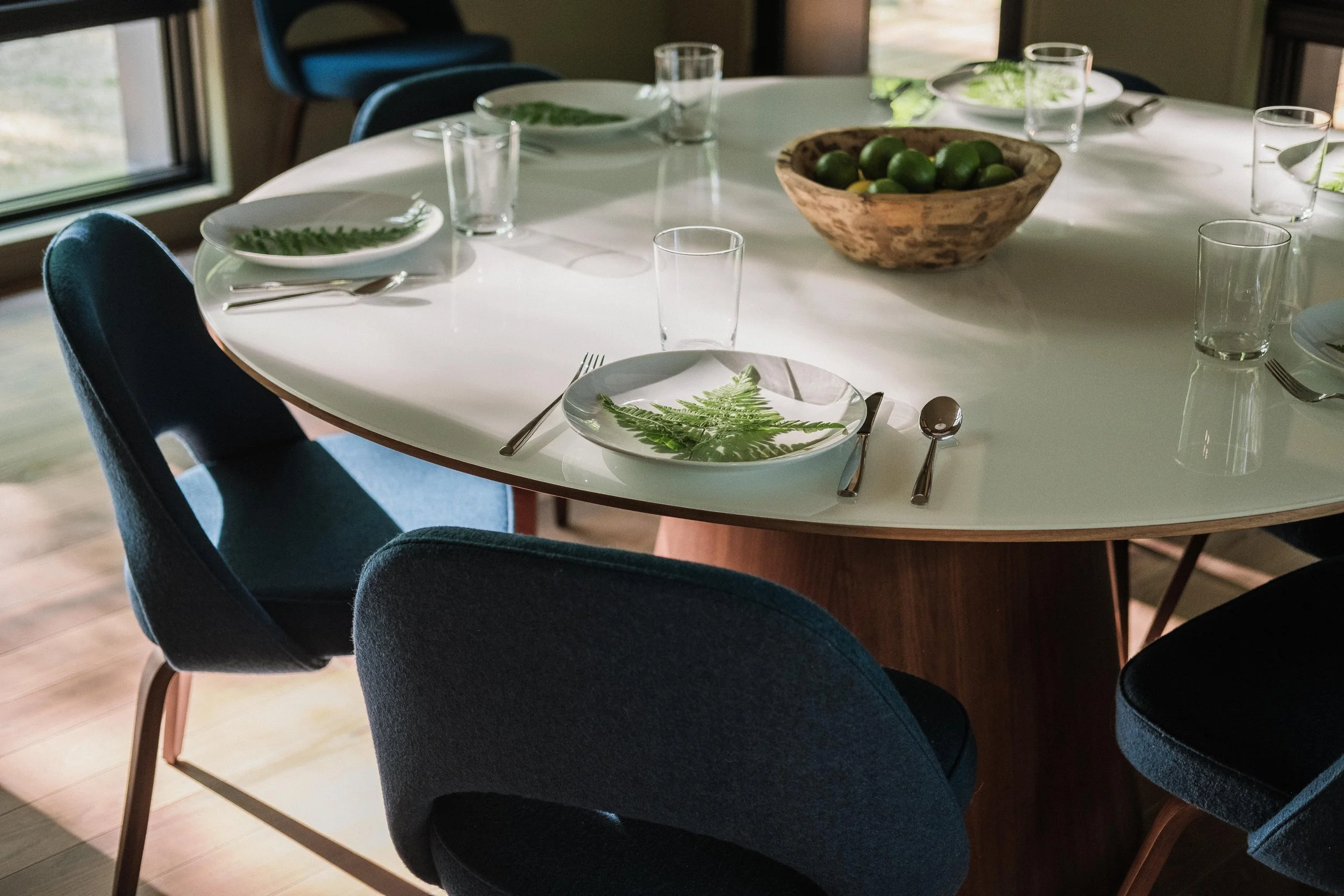 Round dining table set with white plates, silverware, and glasses. Each plate has a green fern leaf. There is a wooden bowl filled with limes at the center of the table. Several blue chairs surround the table, and sunlight streams in through nearby w