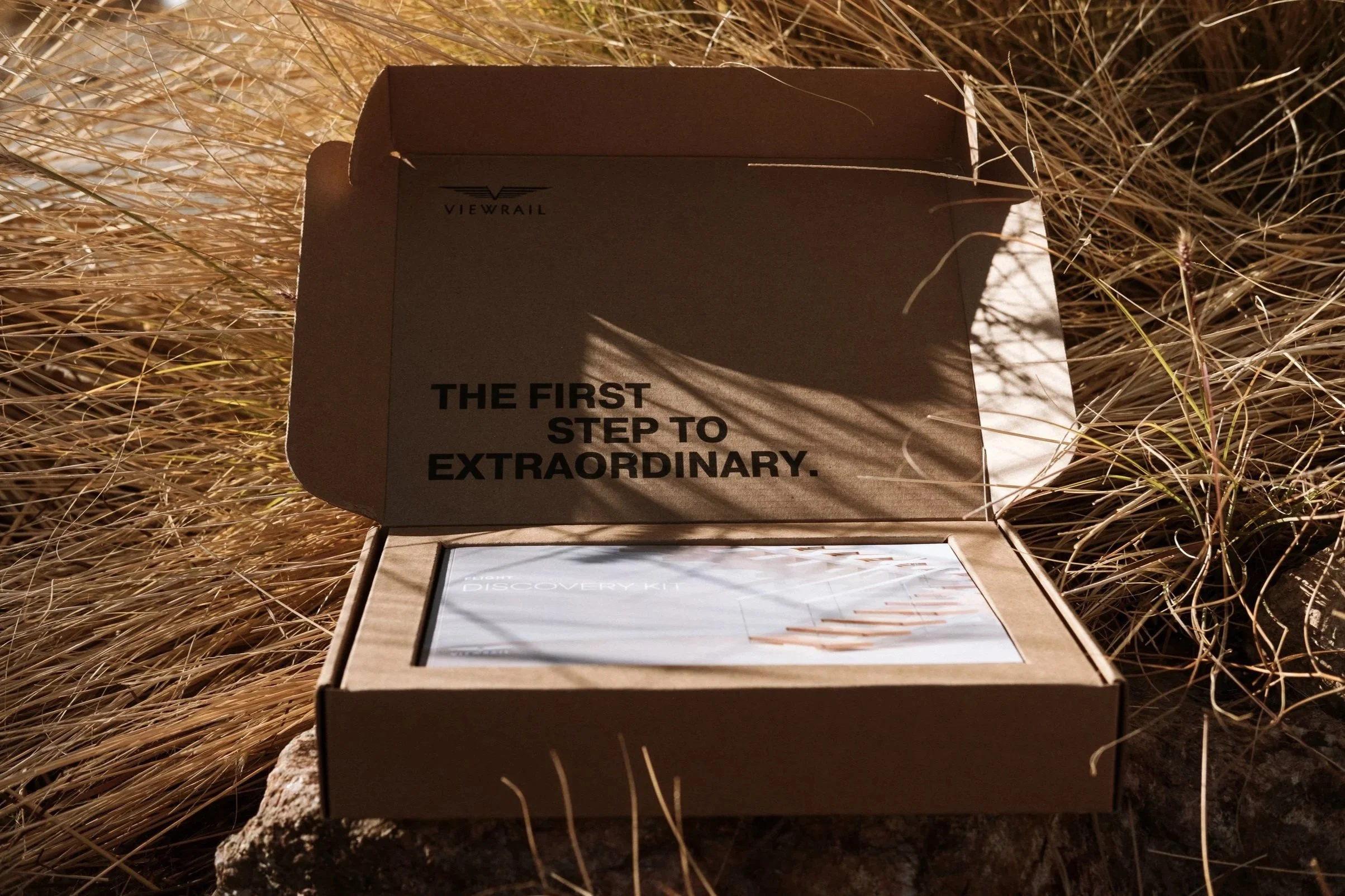 Open cardboard box labeled 'The first step to extraordinary' inside consisting of a discovery kit, placed on the ground surrounded by dry grass.