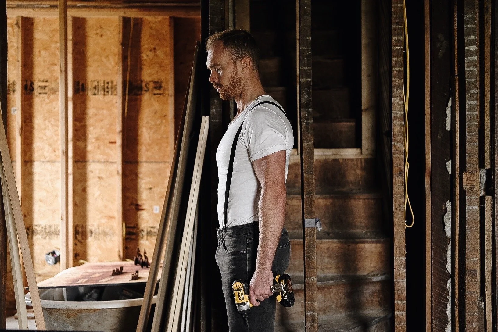 A man in a construction site holds a power drill, standing next to wooden framing and stairs, with a serious expression.