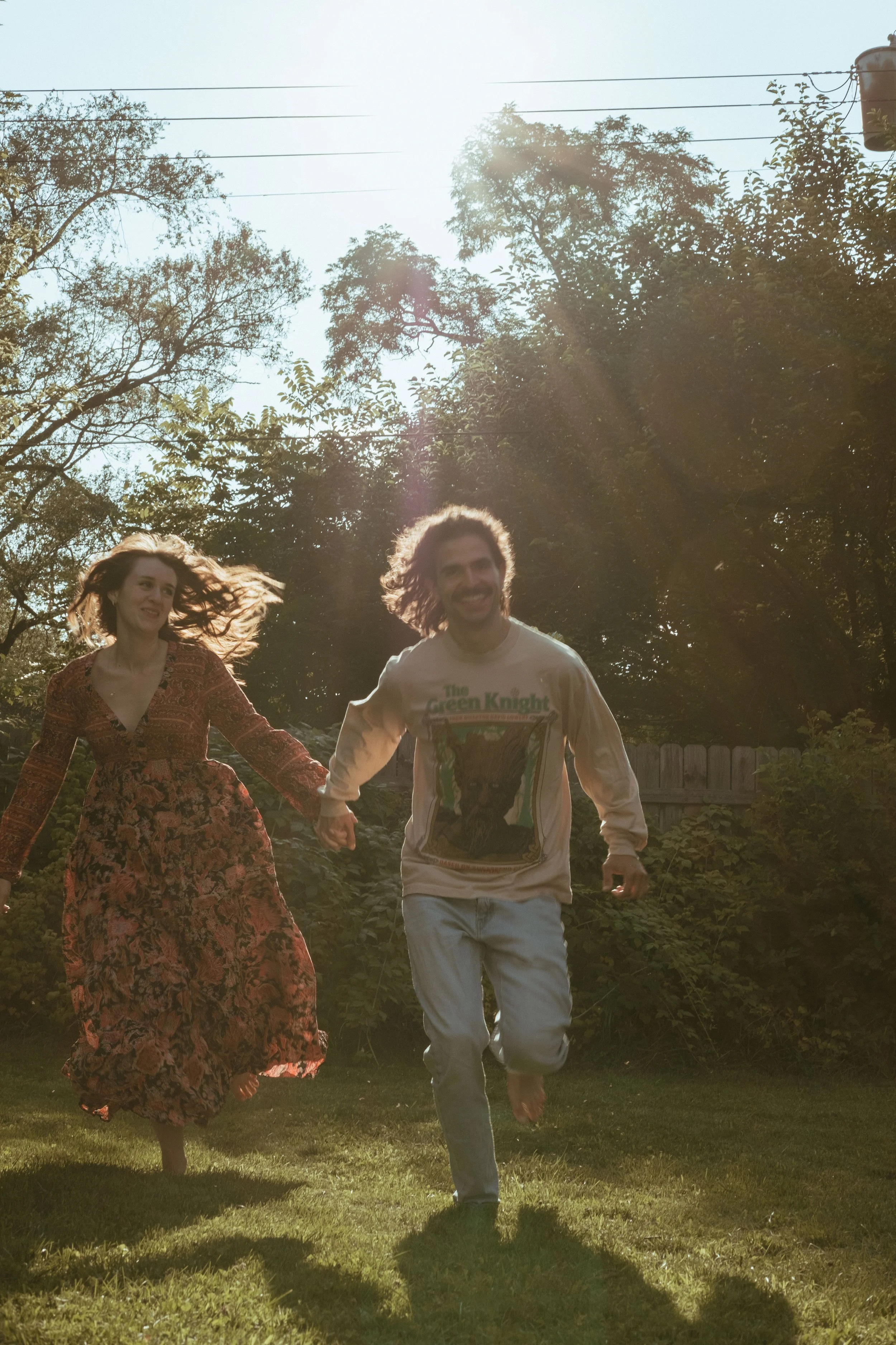 A man and a woman running barefoot on grass in a backyard with trees and a wooden fence, with sunlight shining through the branches.