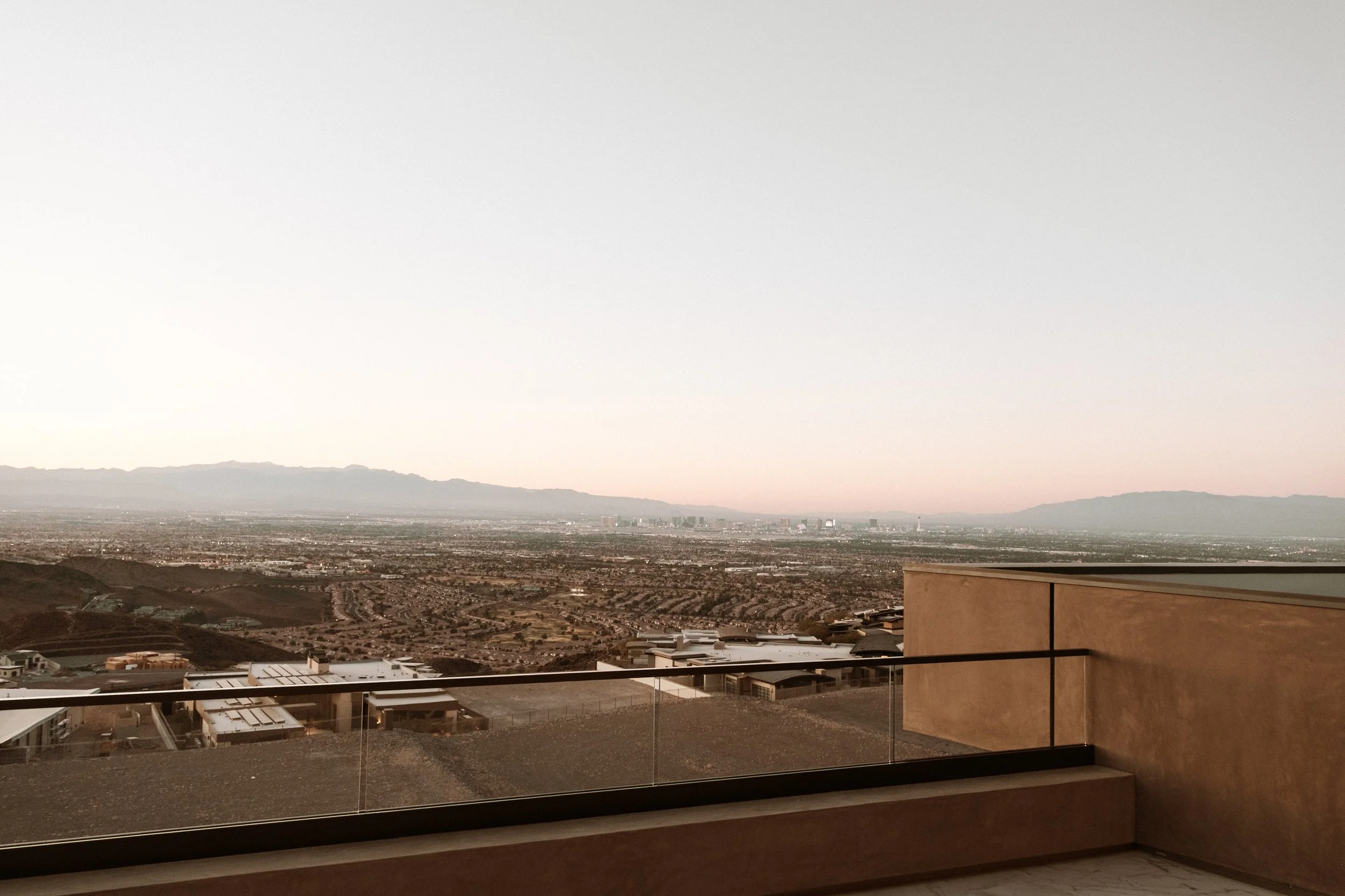 View of a desert landscape with distant mountains, seen from a balcony with a glass railing and a brown wall on the right.