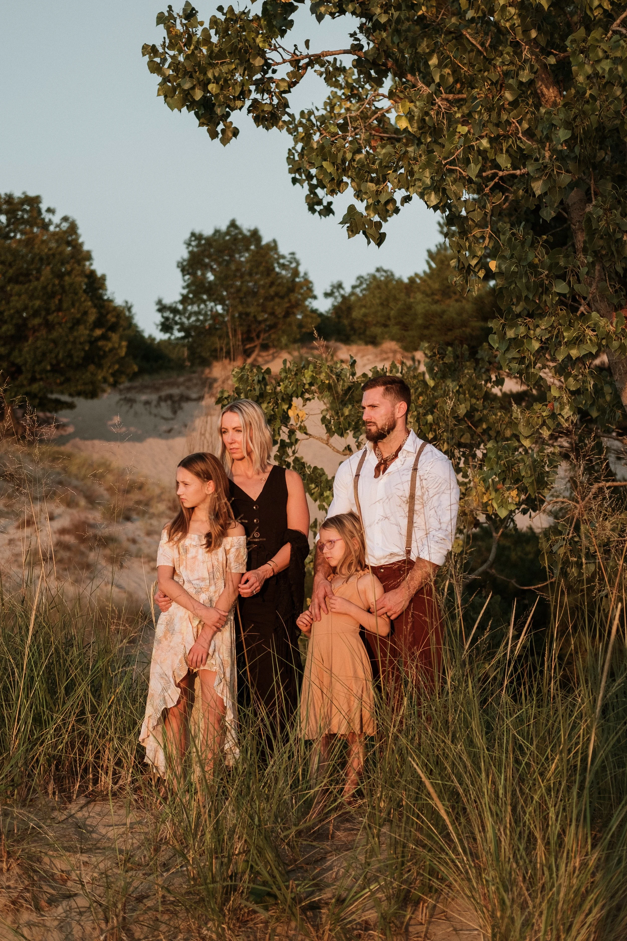Family of four standing together outdoors in a field with tall grass and trees in the background, during sunset.