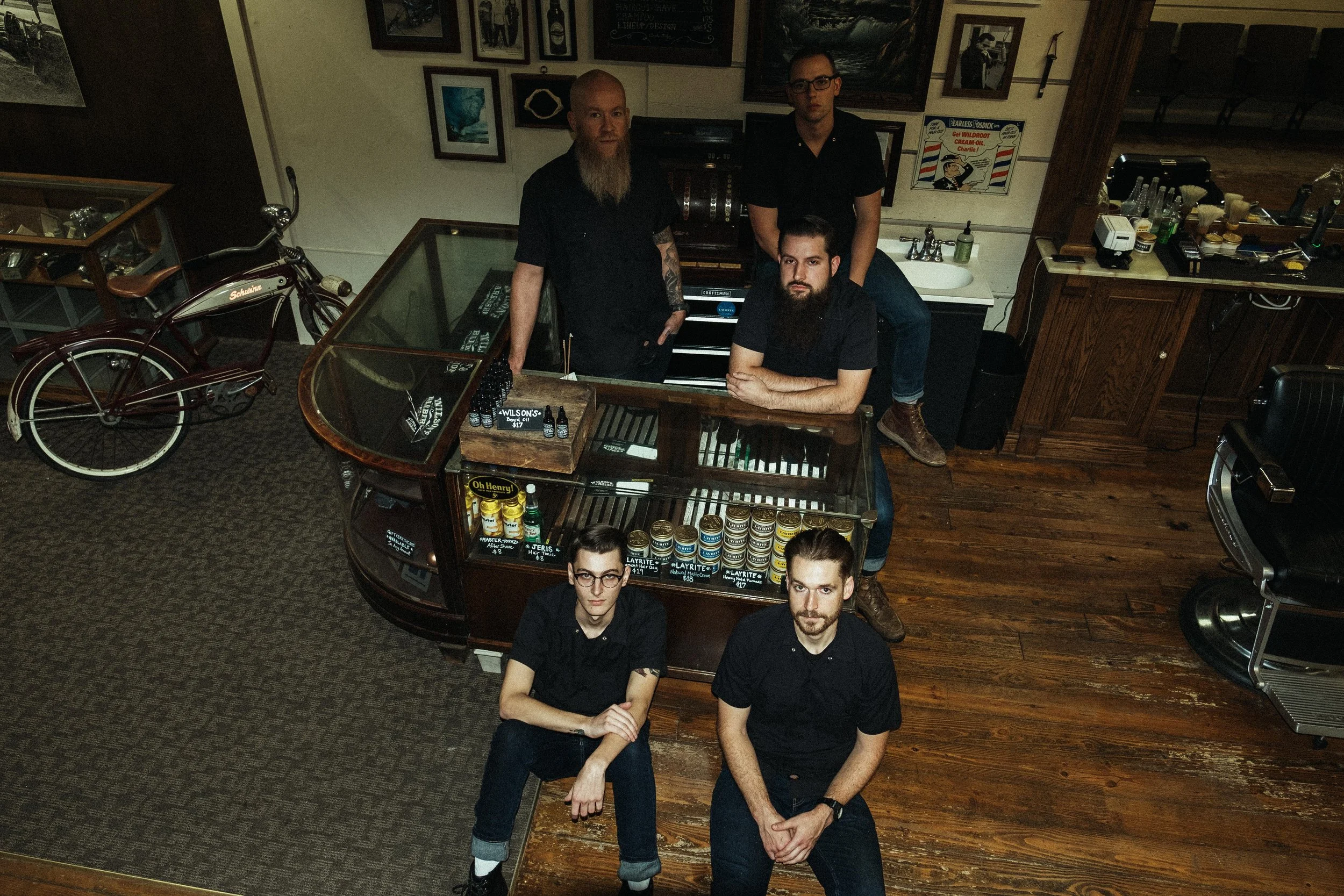Five people in black shirts gathered around a bar inside a vintage-style cafe or bar. Two men are standing behind the counter, one sitting on the bar next to the counter, and two people sitting on the floor in front of the bar. The background has fra