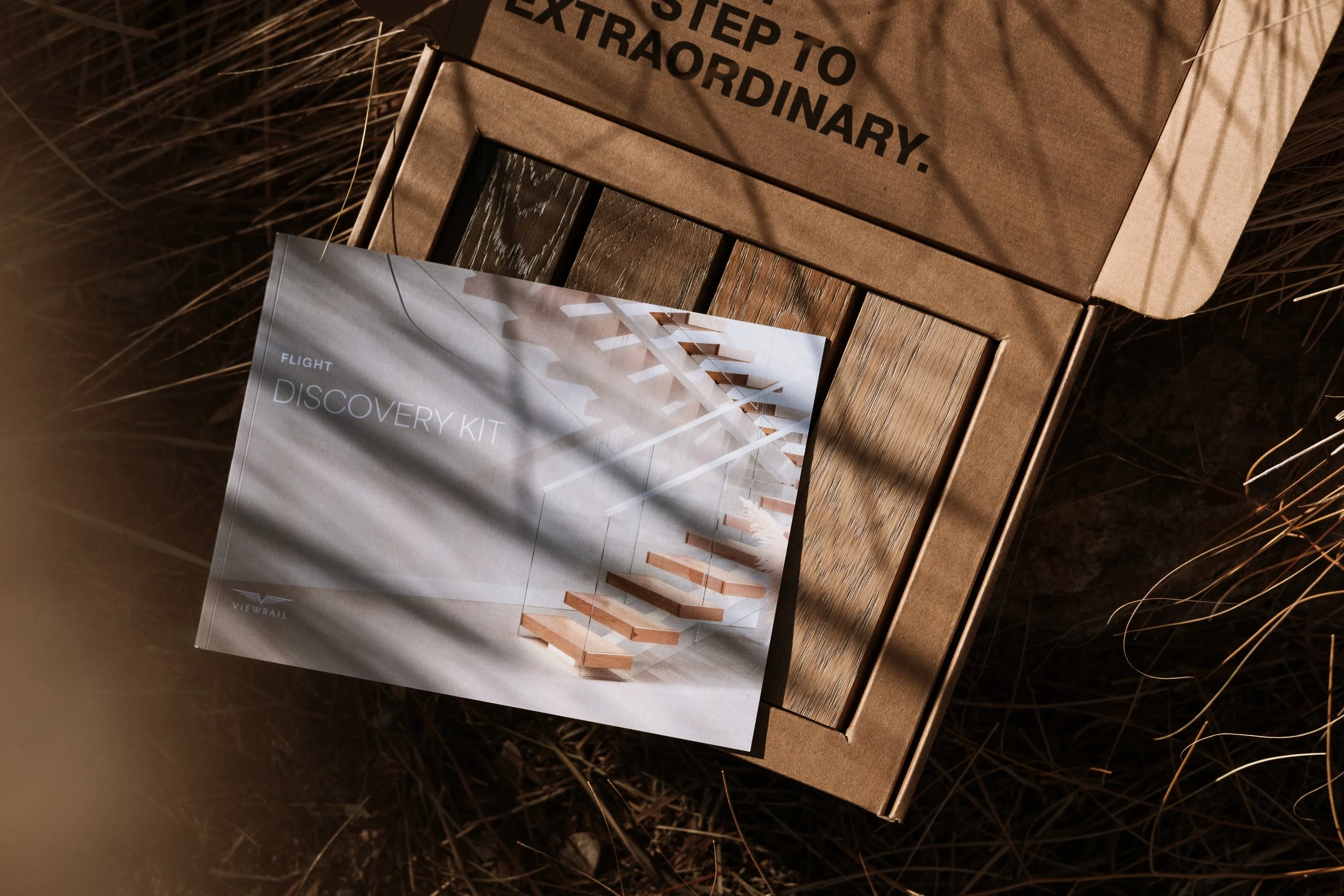 Open cardboard box with a Discovery Kit brochure inside, placed on bamboo or wooden surface outdoors. The brochure features an image of a modern, minimalist staircase design.