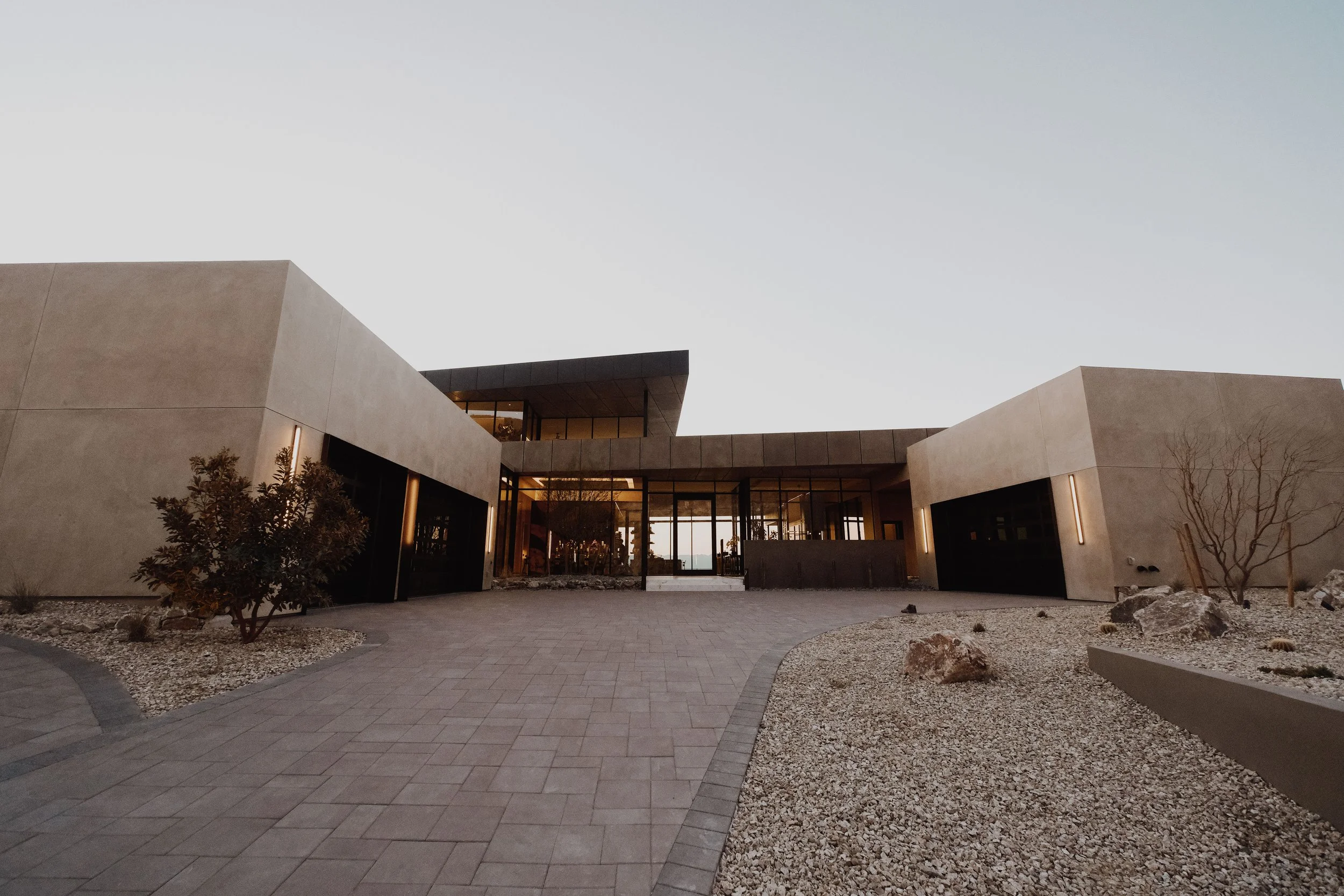 Modern building with beige and black exterior walls, large glass windows, and minimal landscaping with rocks and sparse trees, taken during dusk or dawn.
