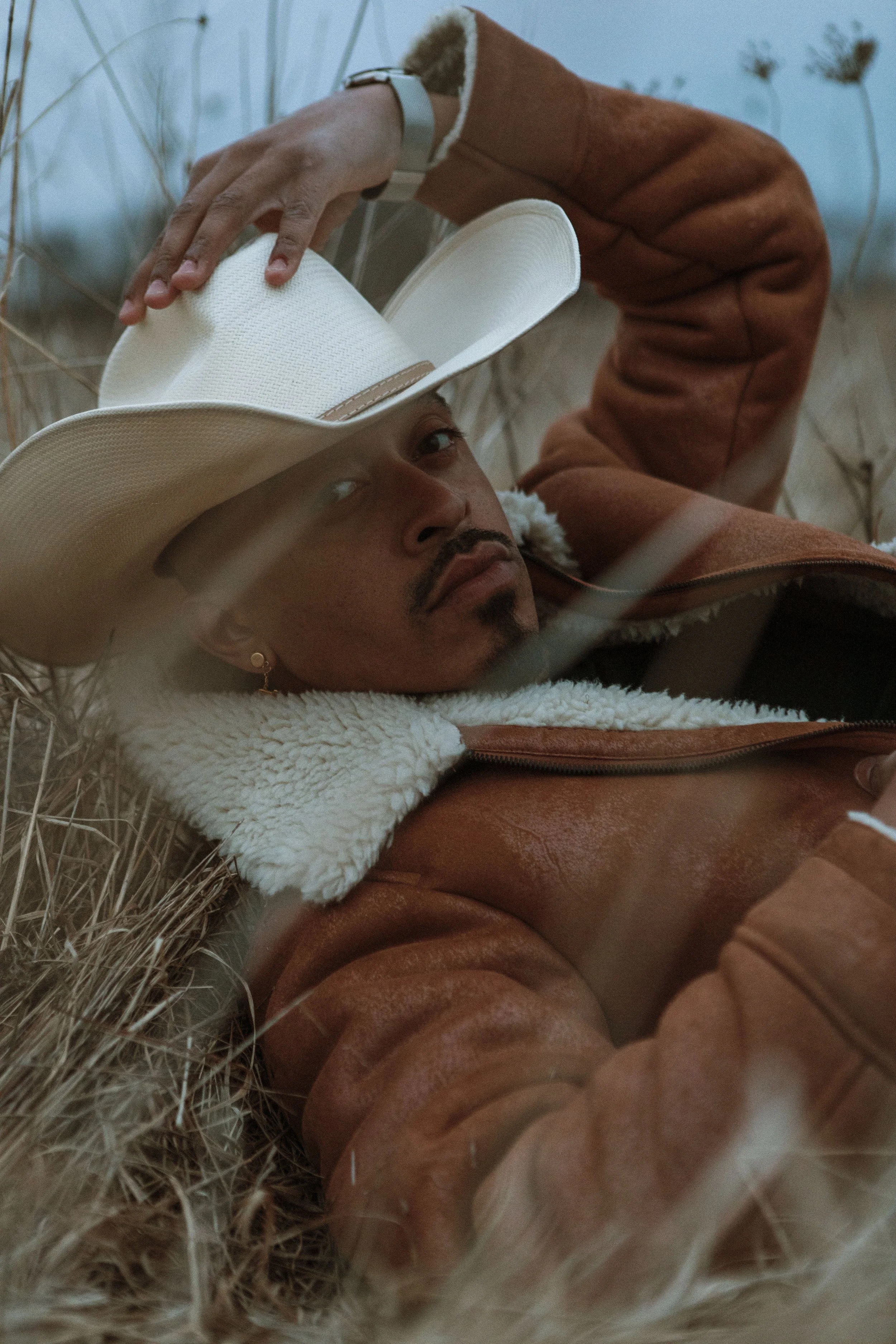 A man lying on a bed of dry grass, wearing a cowboy hat and a brown leather jacket with a shear-lined collar.