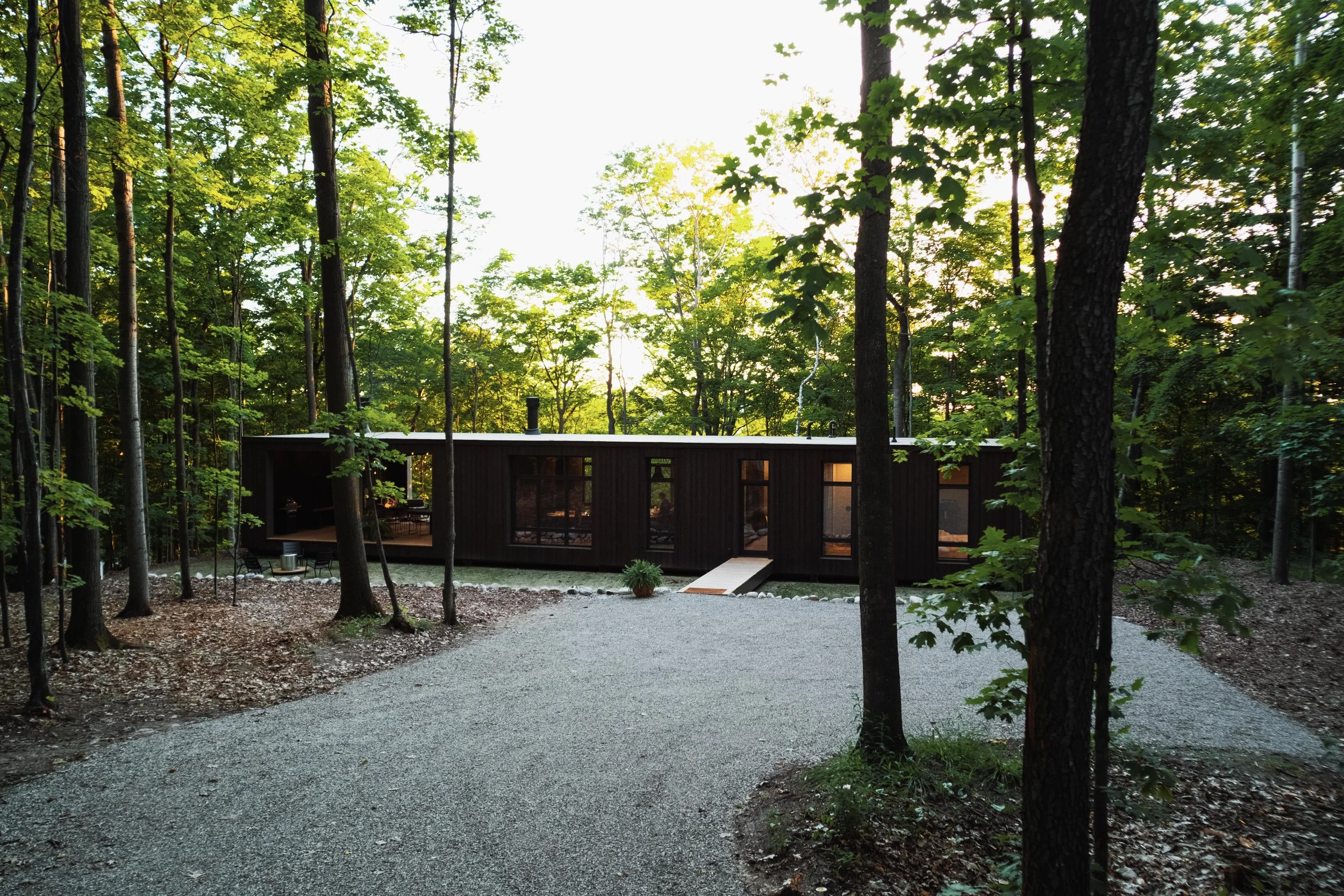 Modern black house in a wooded forest with a gravel driveway and trees surrounding the property, during sunset.