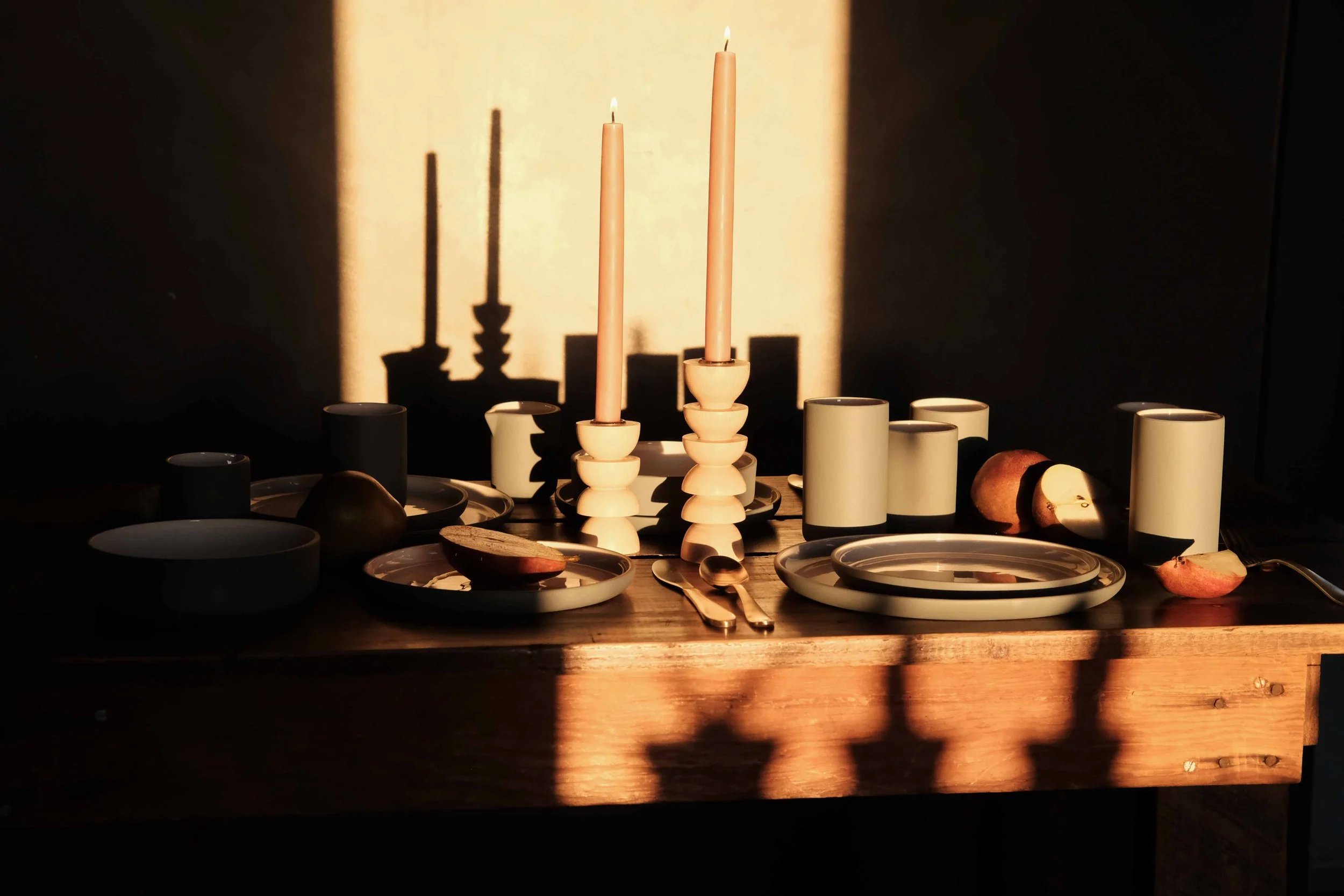A wooden table set for a meal with various dishware, candles, and an apple, illuminated by warm sunlight