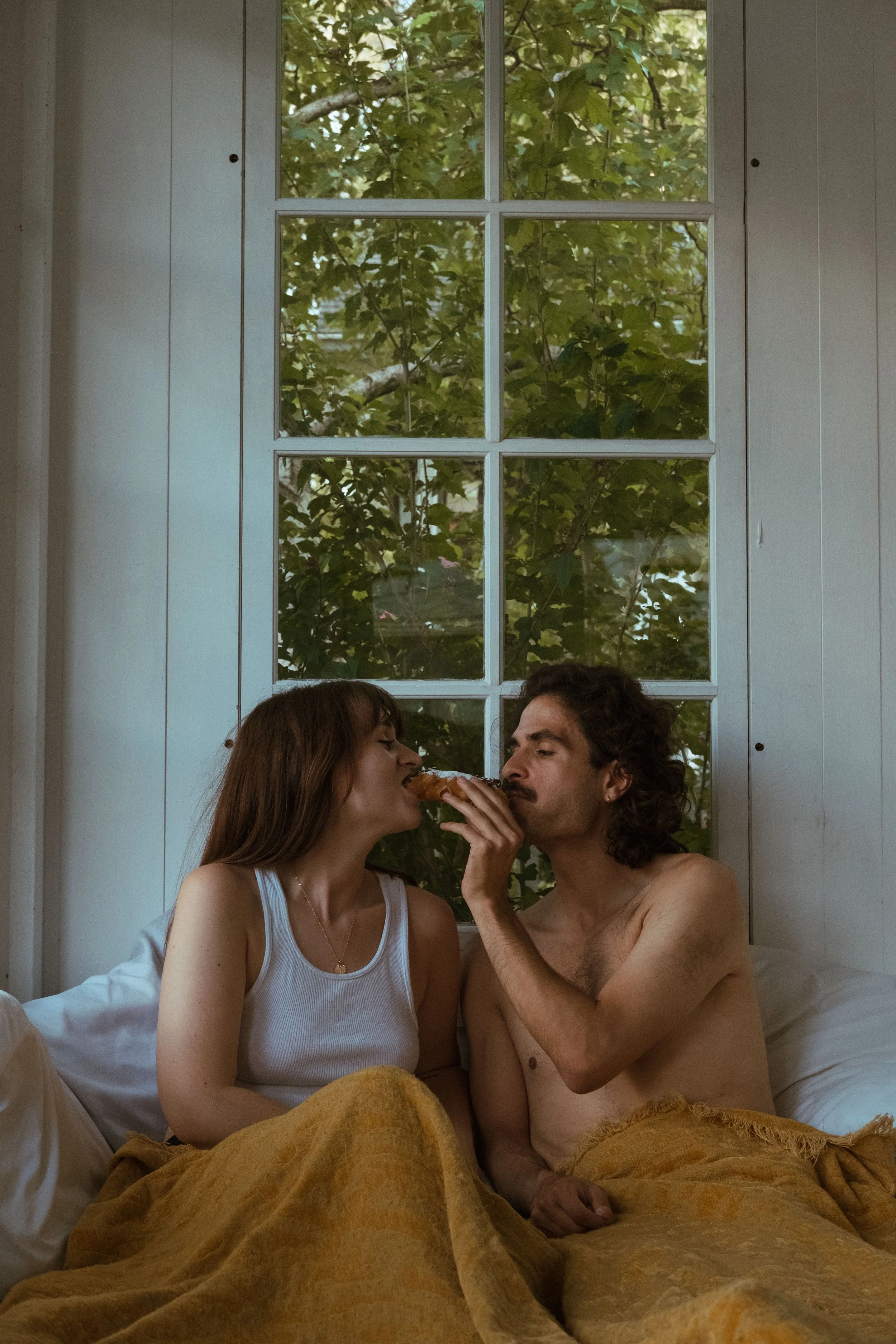 A woman and a shirtless man sitting on a bed between a window and white wooden walls, sharing a slice of pizza, with green foliage visible outside the window.