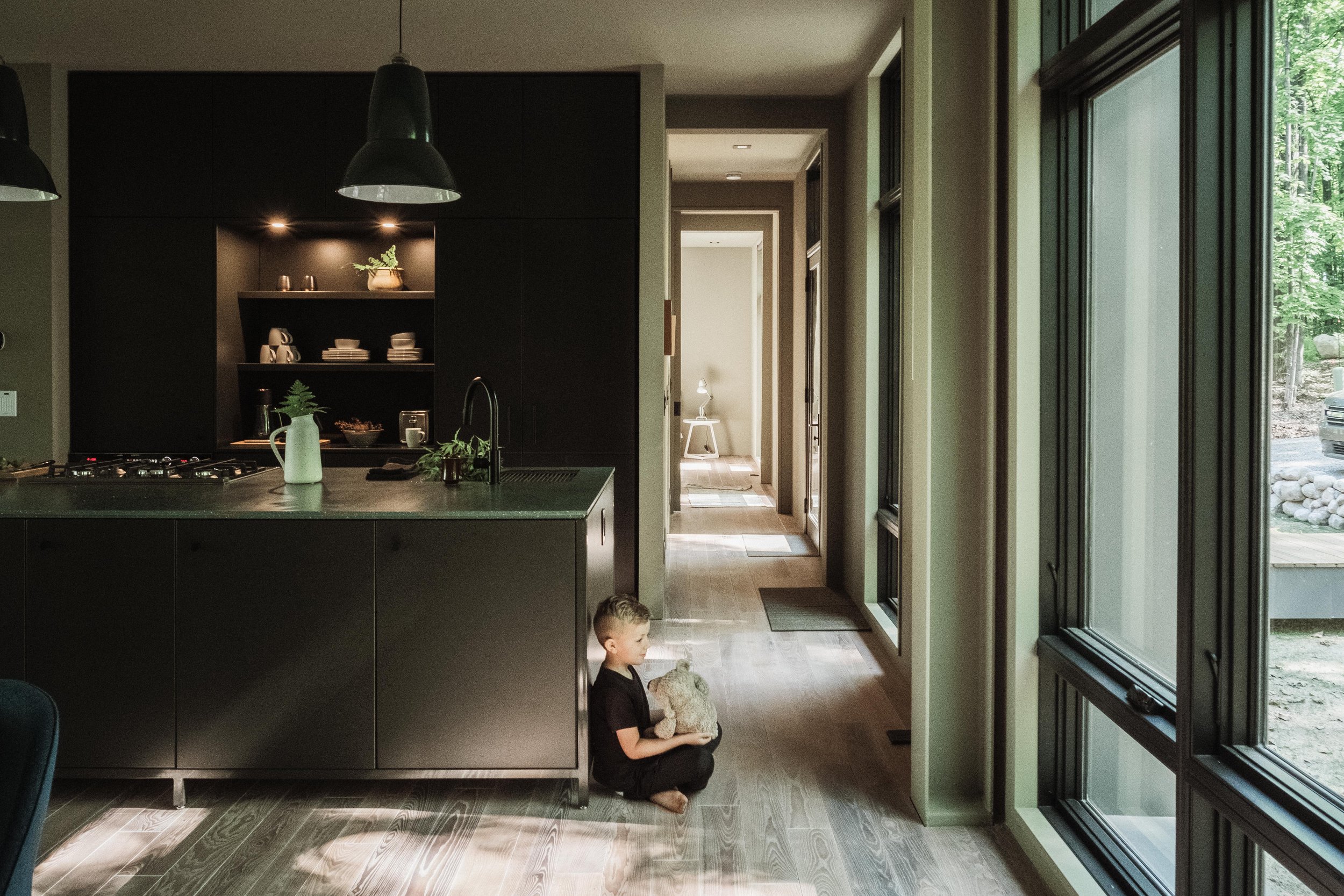A young boy is sitting on the wooden floor in a modern kitchen, holding a stuffed animal, near a large window and a kitchen island with dark cabinets.