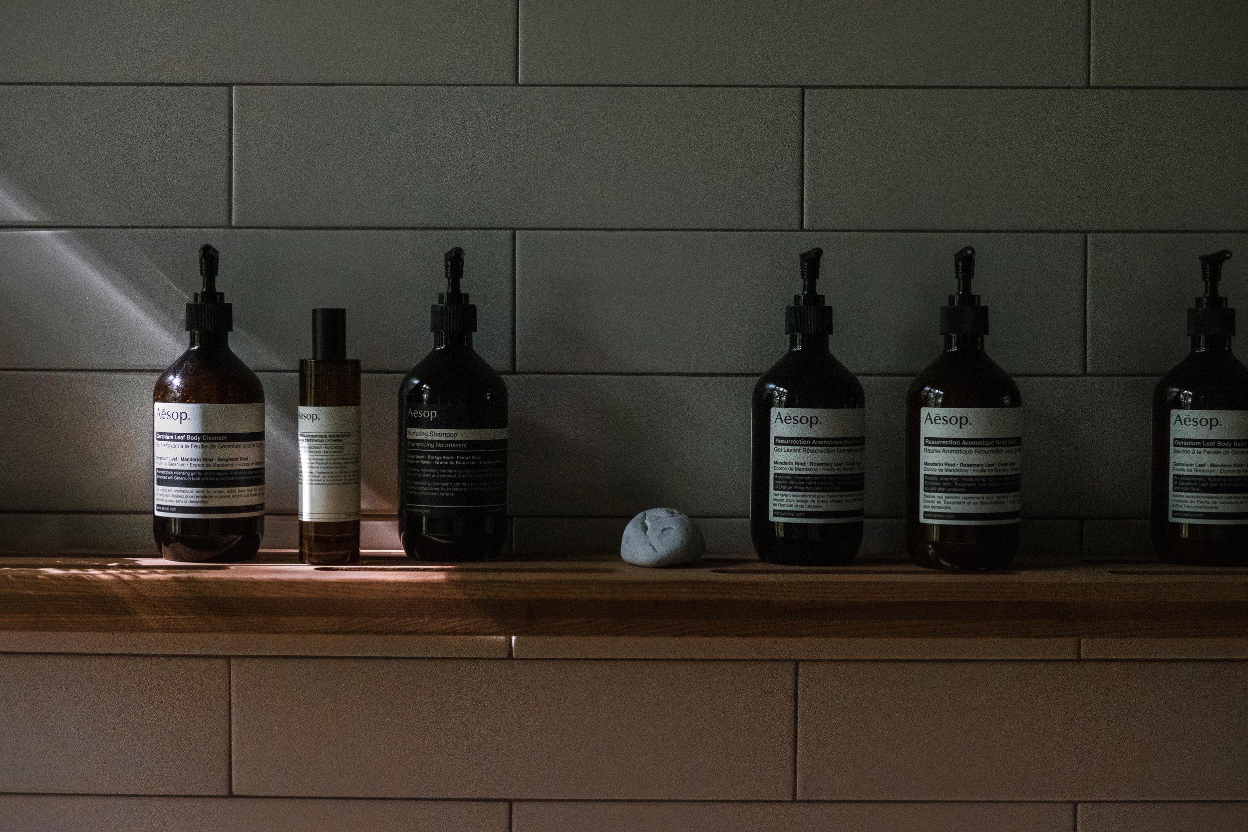 A row of black and brown bottles with pump dispensers and labels on a wooden shelf, accompanied by a small white stone, set against a tiled wall.