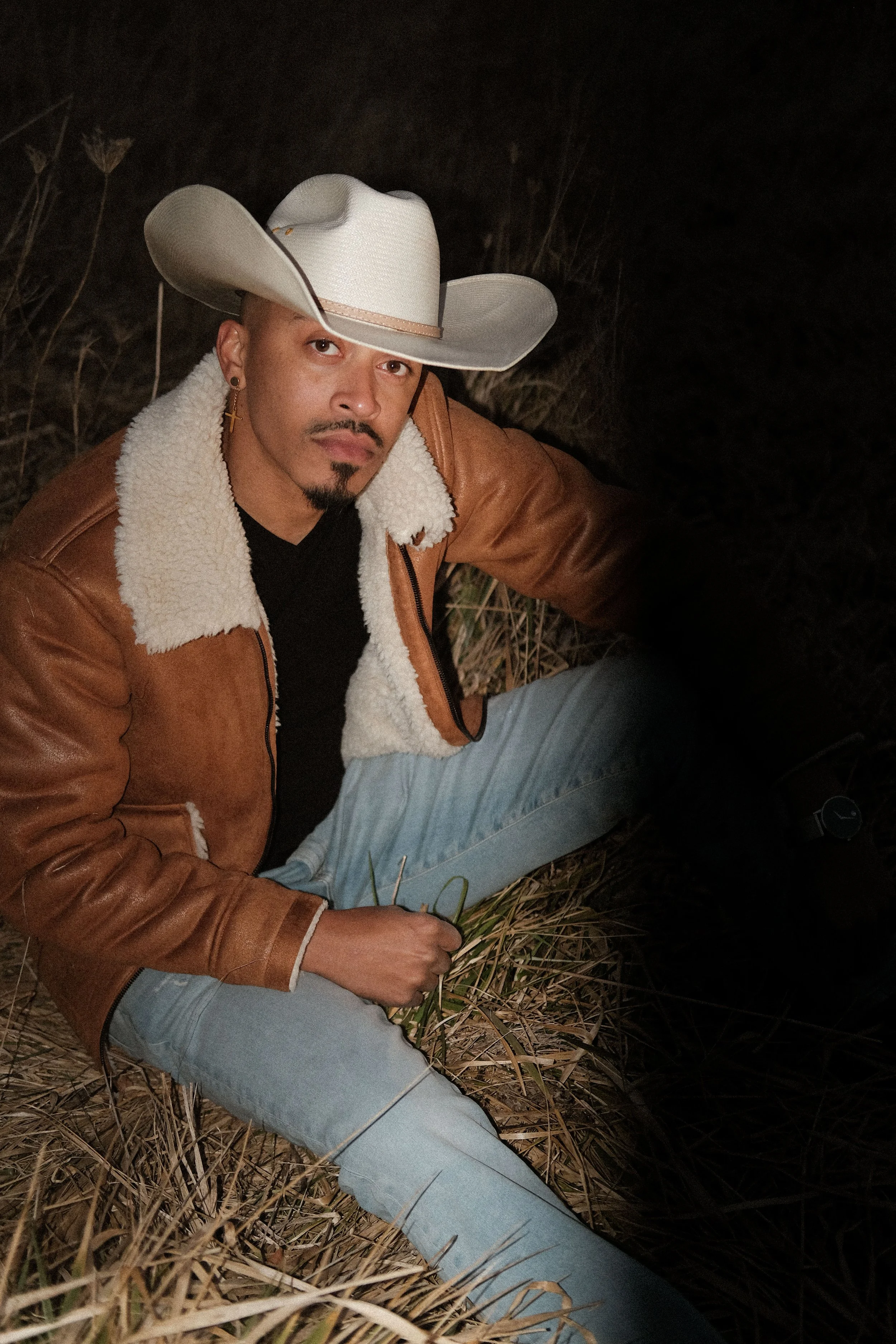 A man sitting on the ground in a field at night, wearing a cowboy hat, a brown leather jacket with a shear lining, a black shirt, and light blue jeans.