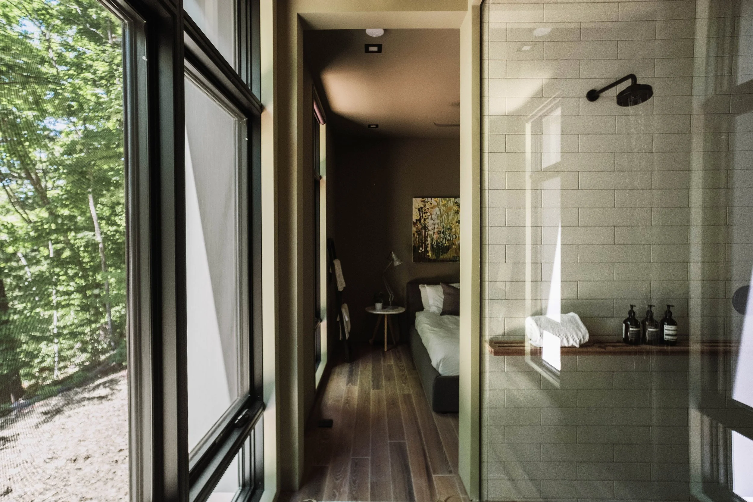 Interior view of a modern bedroom and bathroom with large windows showing green trees outside, a bed with pillows, a side table with a lamp, and bathroom elements including a shower with a black showerhead and bottles, a towel, and tiles.
