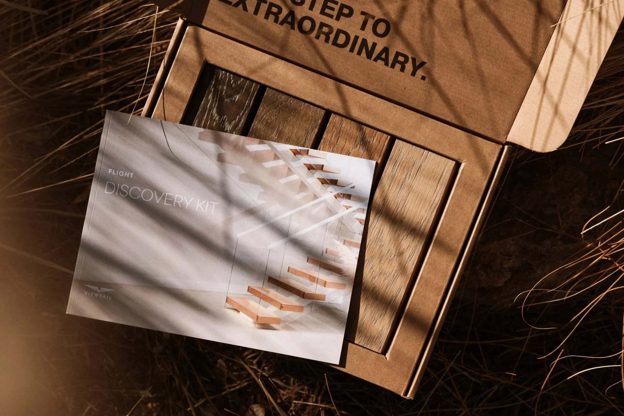 Open cardboard box labeled 'STOP TO EXTRAORDINARY'. Inside the box is a booklet titled 'FLIGHT DISCOVERY KIT' showing a modern staircase on its cover, placed on dry grass and casting shadows.