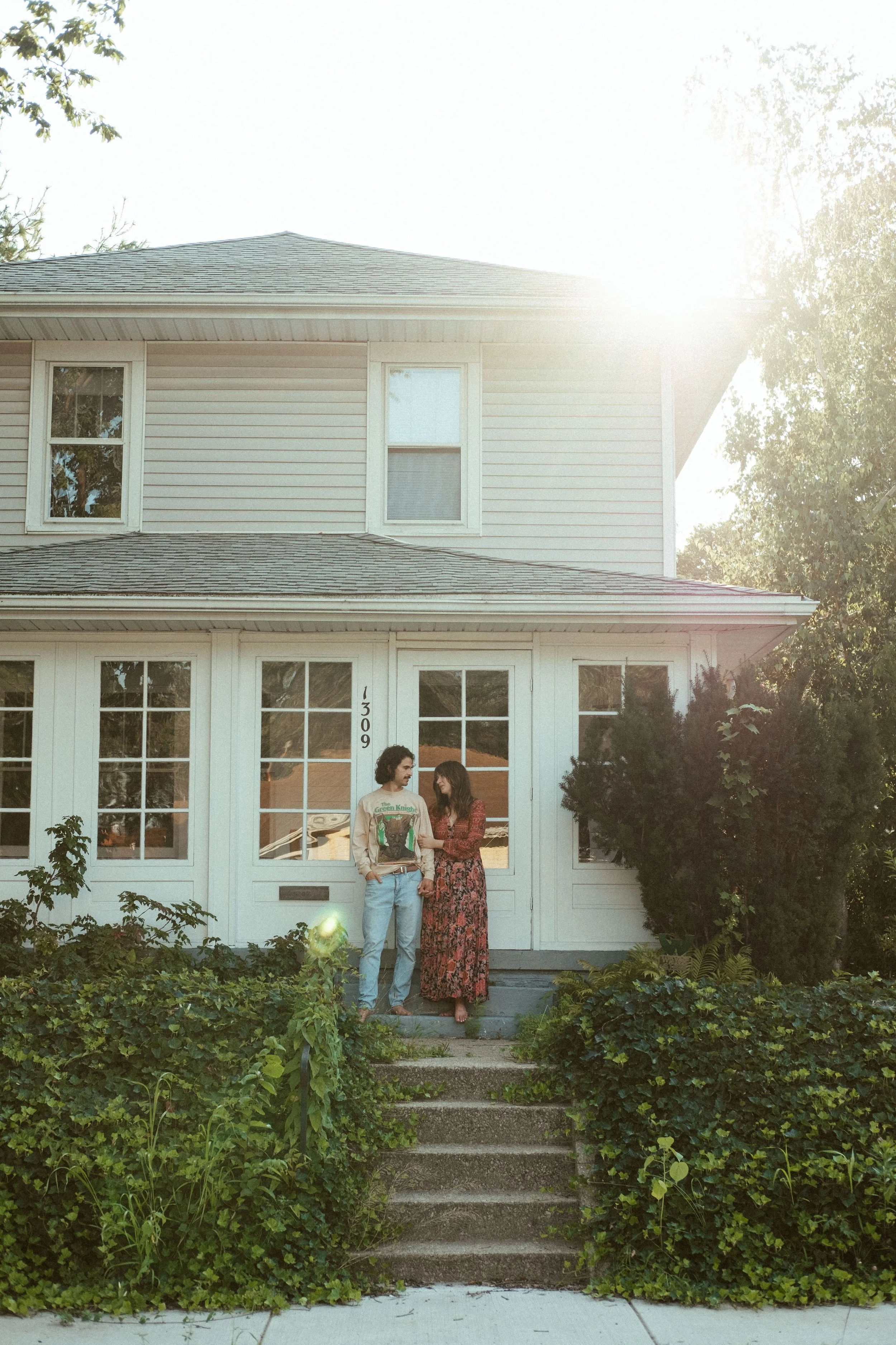 Two people standing on the front steps of a house, holding hands, with sunlight in the background.