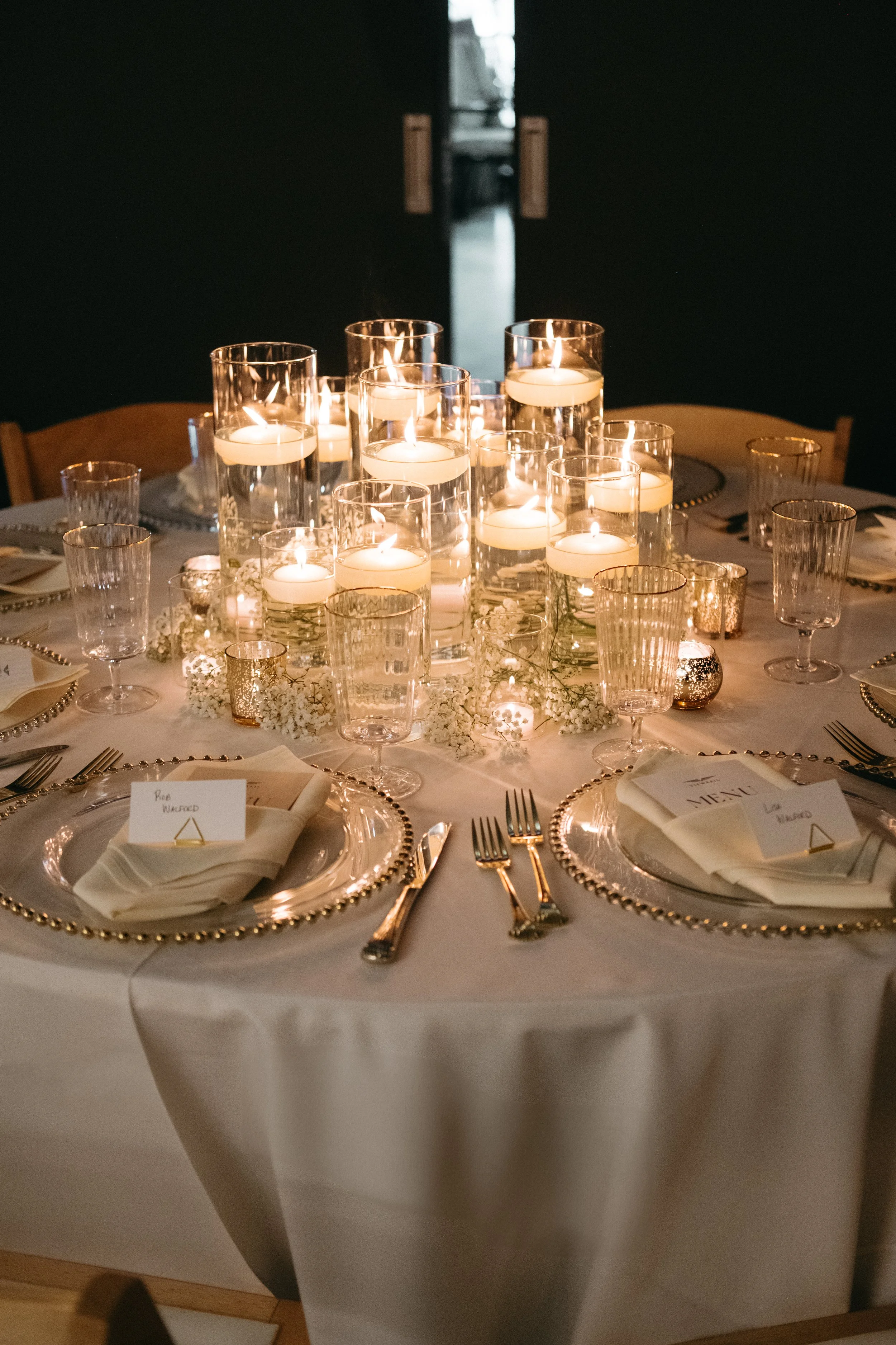 Elegant round dining table set for an event with a white tablecloth, multiple lit candles in glass holders as a centerpiece, and place settings with silverware, napkins, and menu cards.