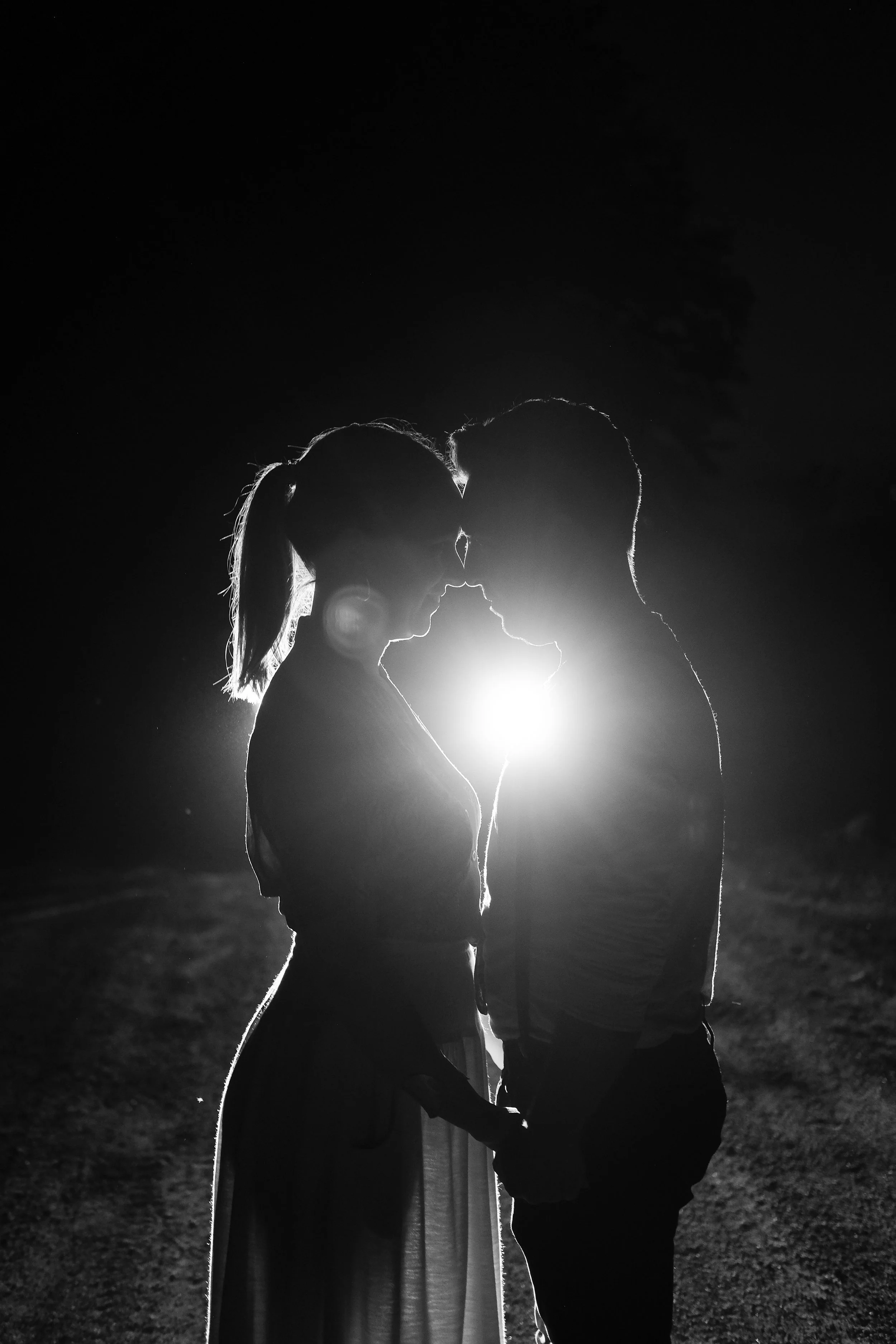 Silhouette of a couple holding hands and facing each other at night with backlighting from a bright source behind them.