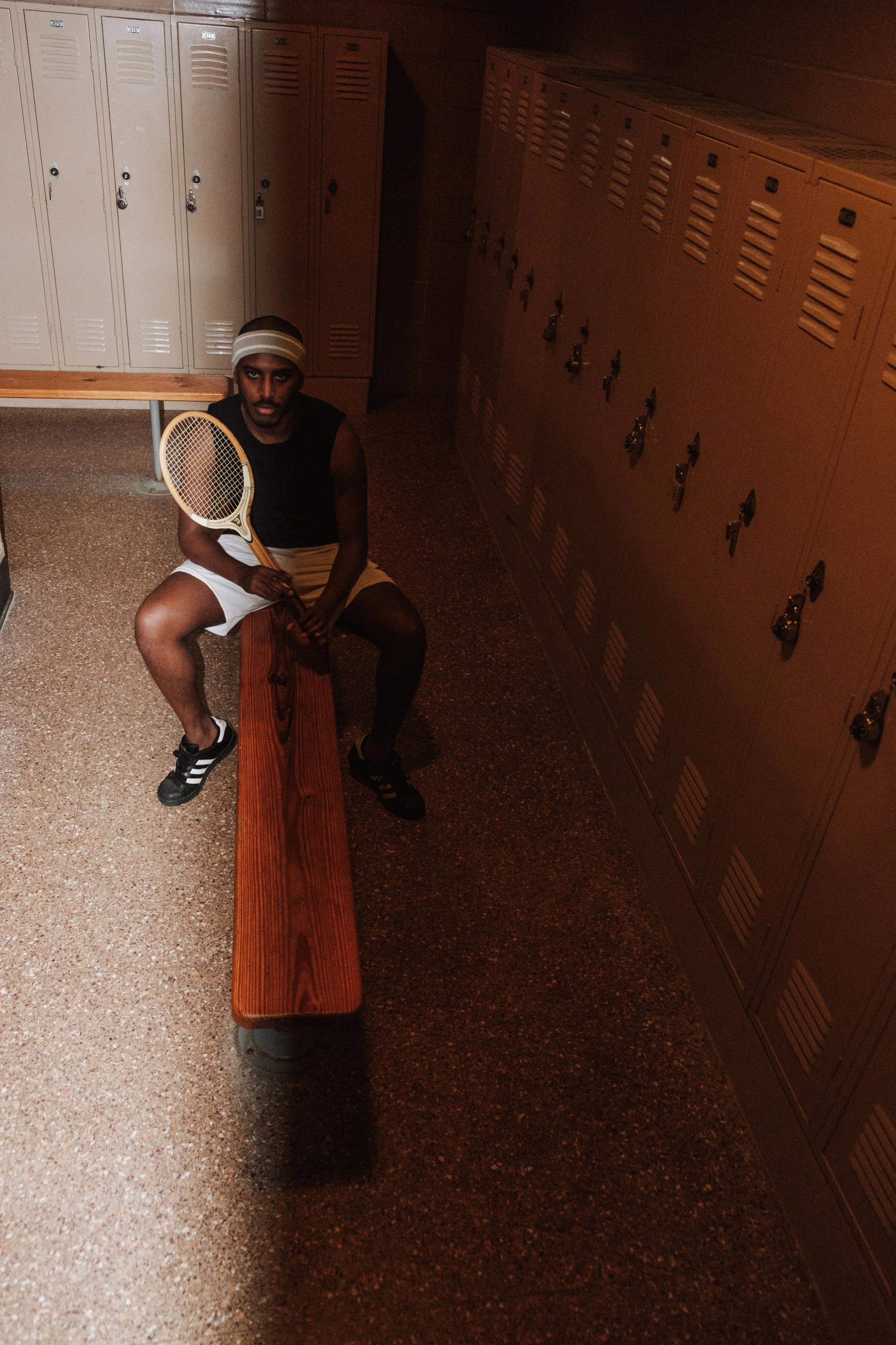 A young man sitting on a wooden bench in a locker room, holding a tennis racket, wearing a headband, black sleeveless shirt, white shorts, and black sneakers.