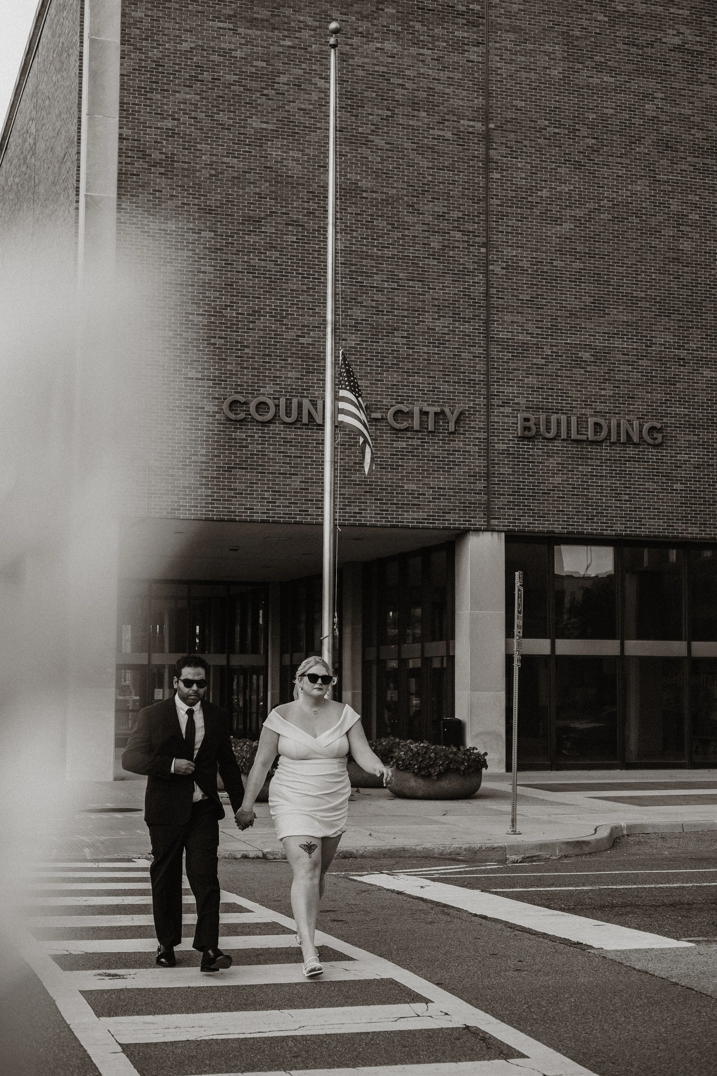 A man and woman holding hands crossing the street in front of a county city building with flags, in black and white.