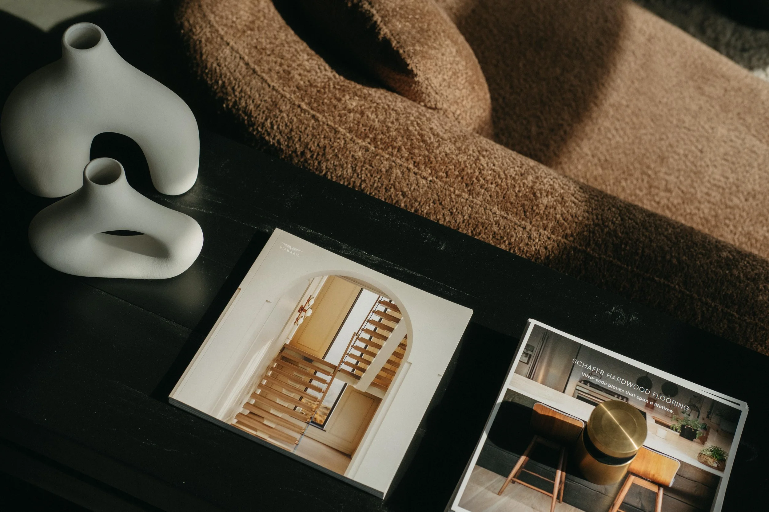 Two white modern ceramic vases with abstract shapes on a black surface, partially covered by a brown textured fabric, next to catalogs showing interior design images and flooring options.