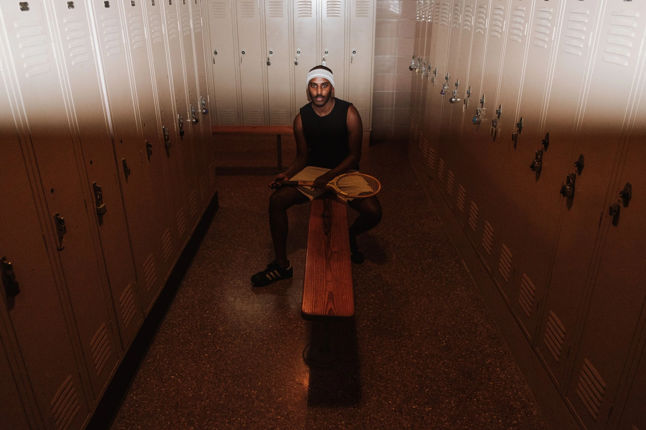 A person sitting on a wooden bench in a locker room, holding a tennis racket and a notebook, wearing black athletic wear, black sneakers, and a white headband.