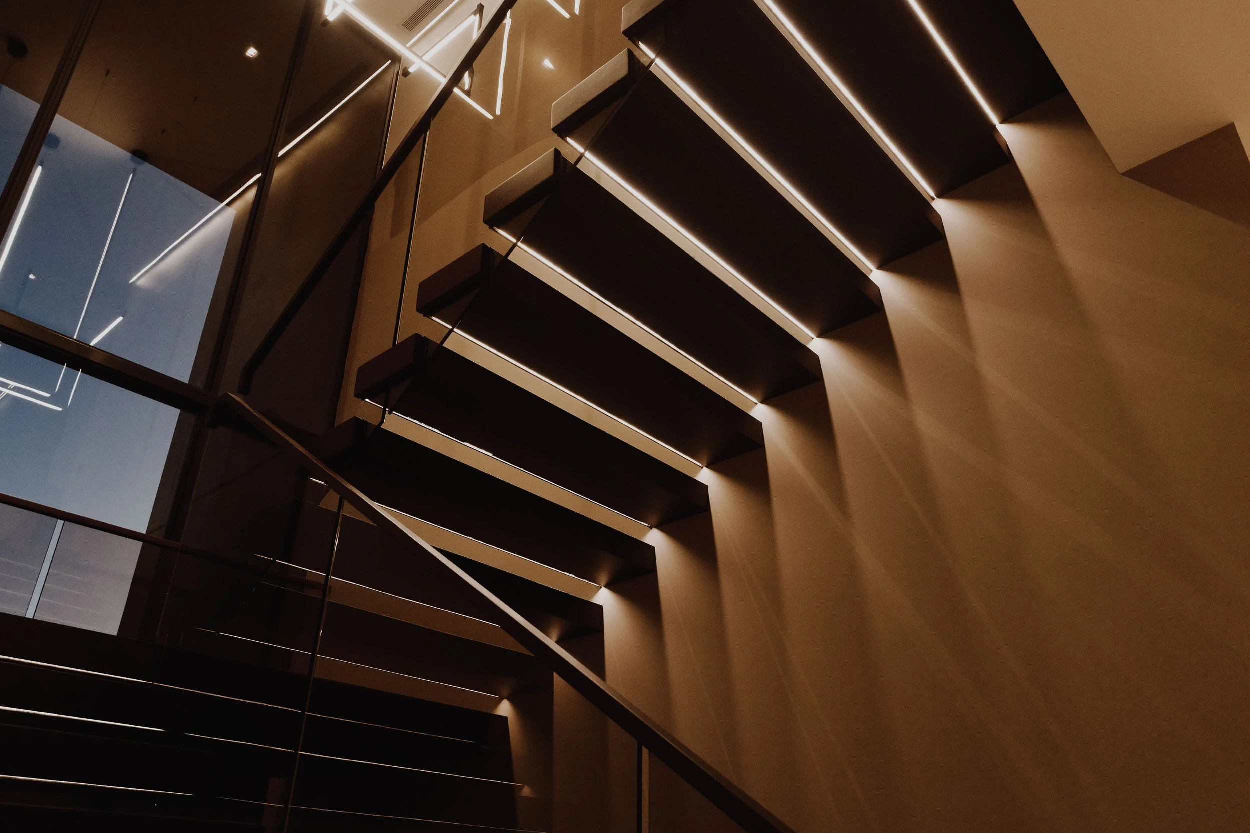Modern interior staircase with horizontal black steps and glass railing, illuminated by linear lighting on the wall and ceiling, viewed from below.