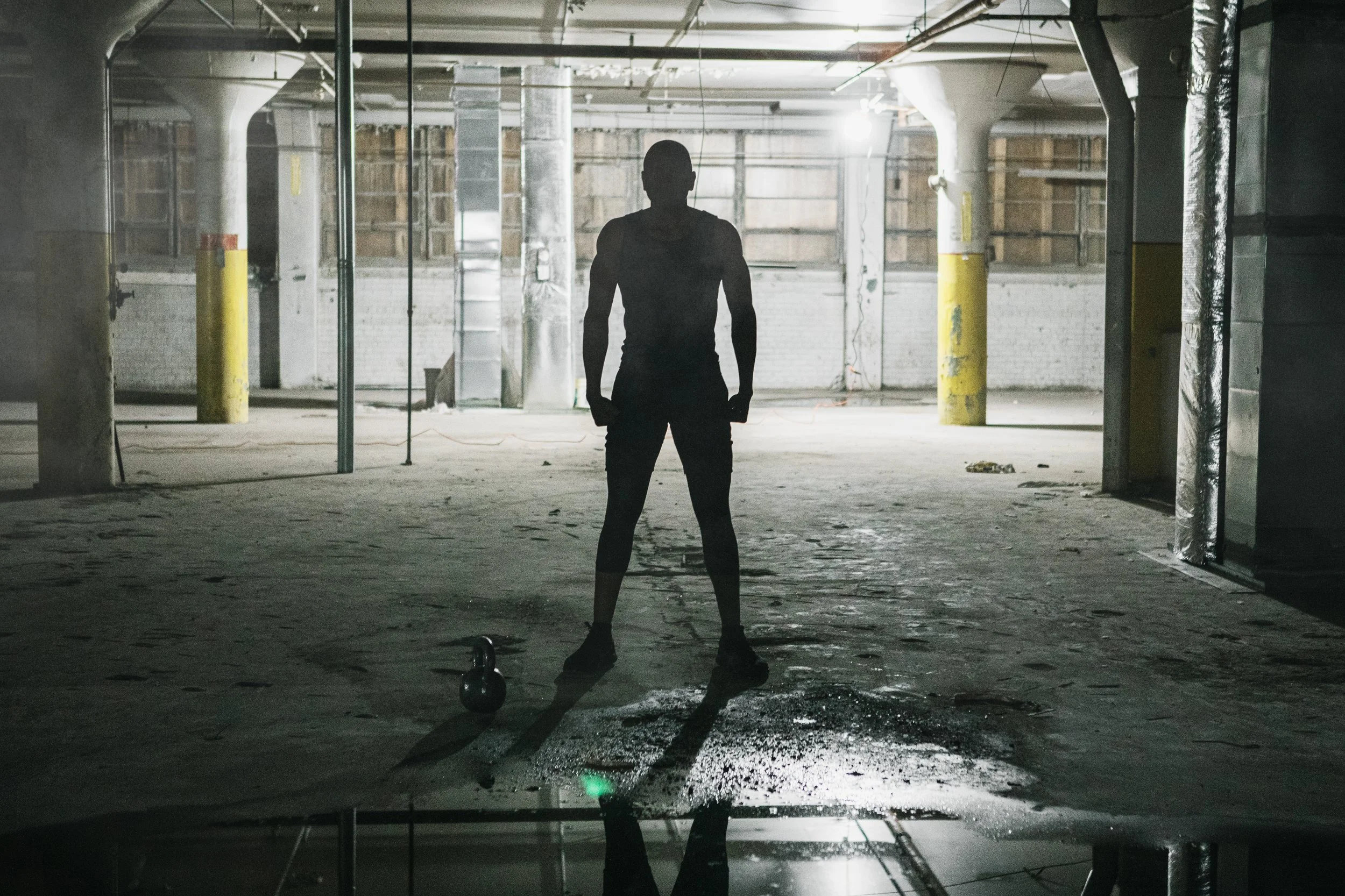A silhouette of a man standing in an empty, industrial parking garage with a kettlebell on the ground nearby and bright light behind him.