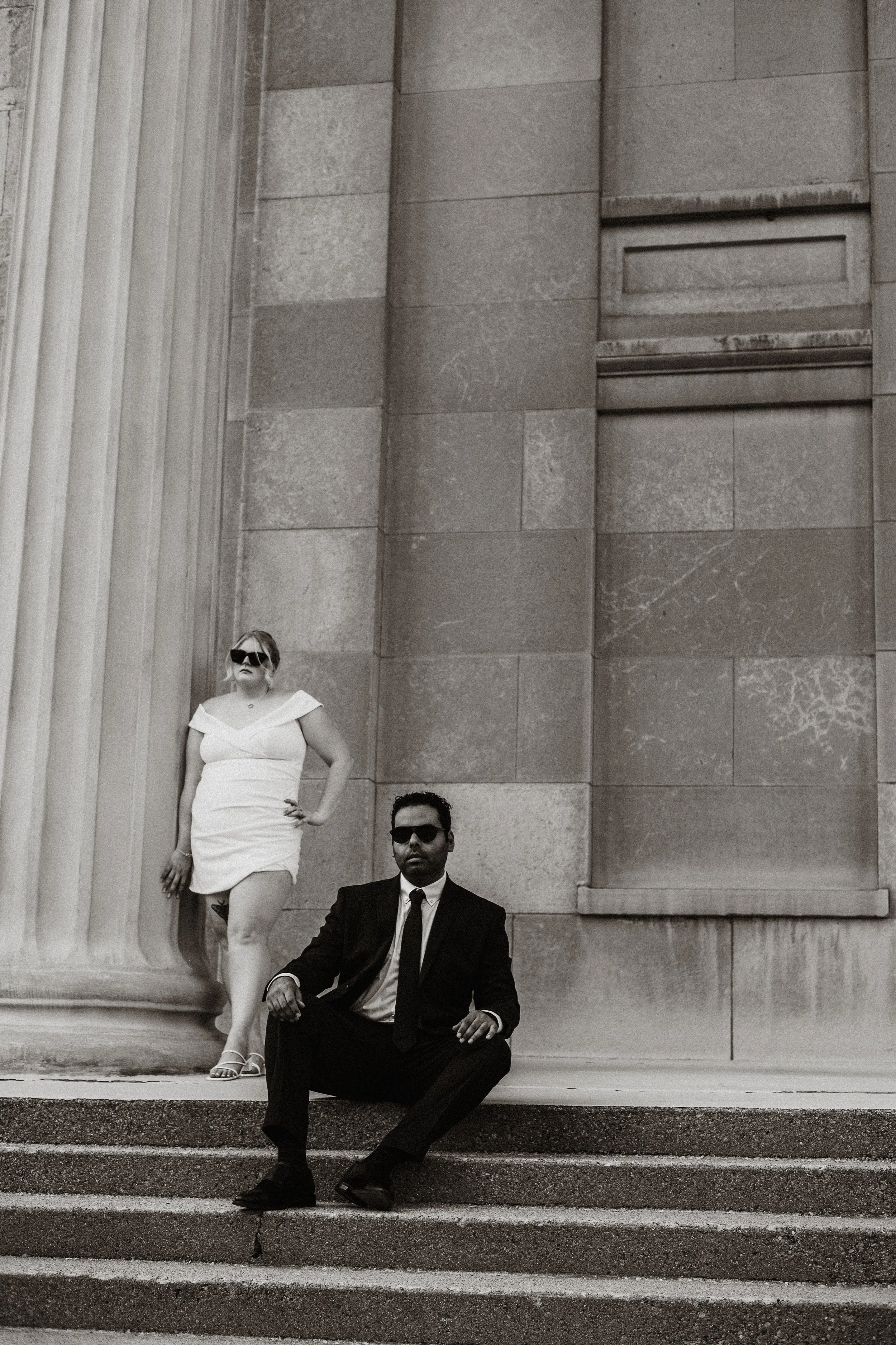 A black and white photo of a woman in a white dress and sunglasses standing next to a seated man in a black suit and sunglasses on the steps of a large building with stone columns and walls.