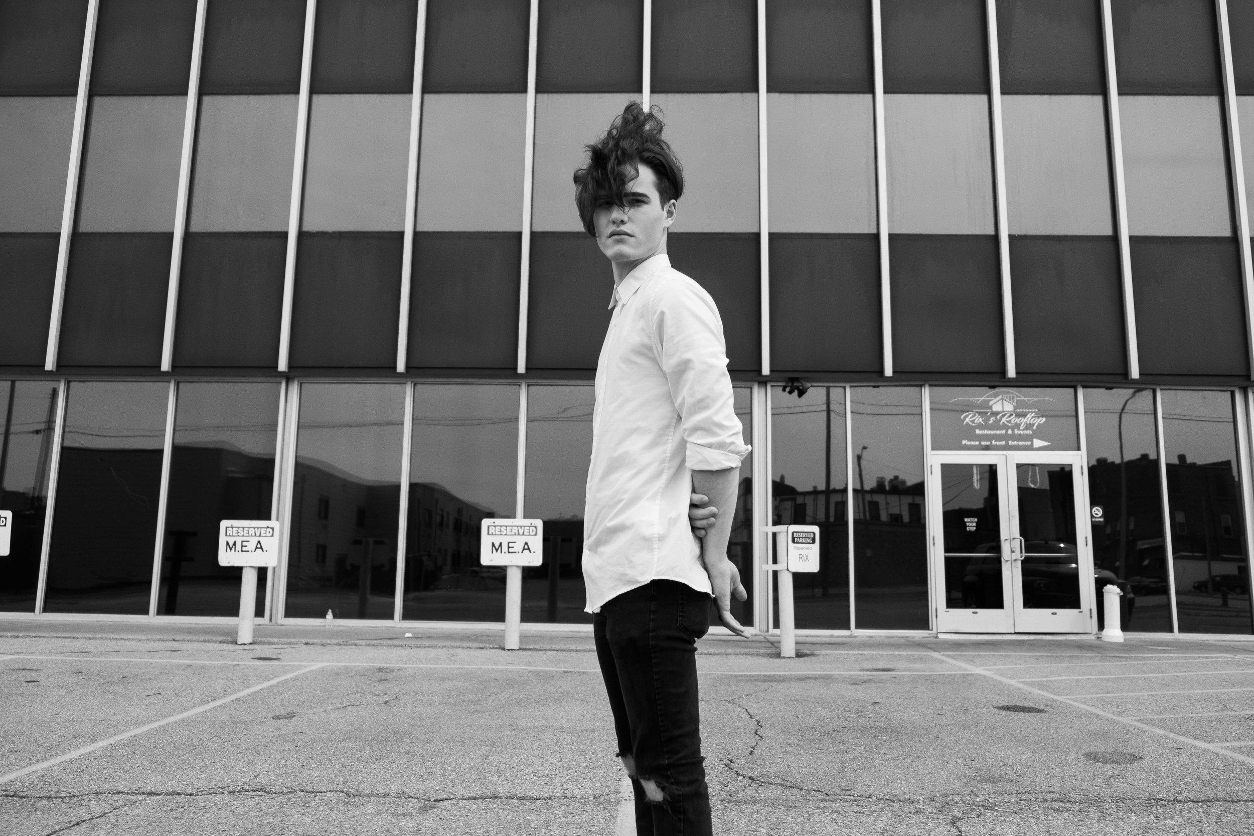 A young person with styled hair, wearing a white shirt and ripped jeans, standing in an empty parking lot in front of a modern building with glass windows.