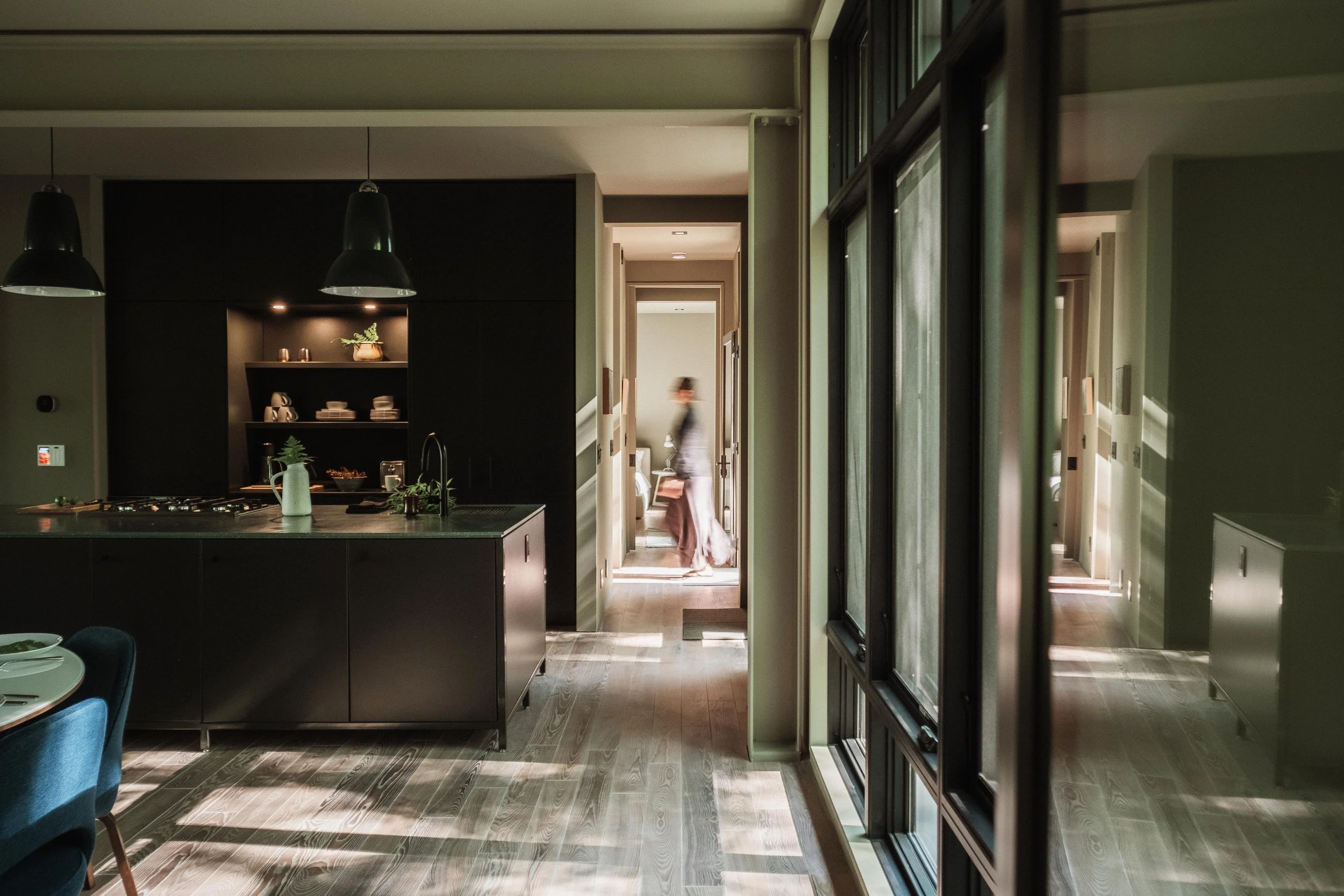 Interior view of a modern home with an open kitchen, black cabinets, a kitchen island, and a hallway with sunlight streaming in. A person is walking away in the background.