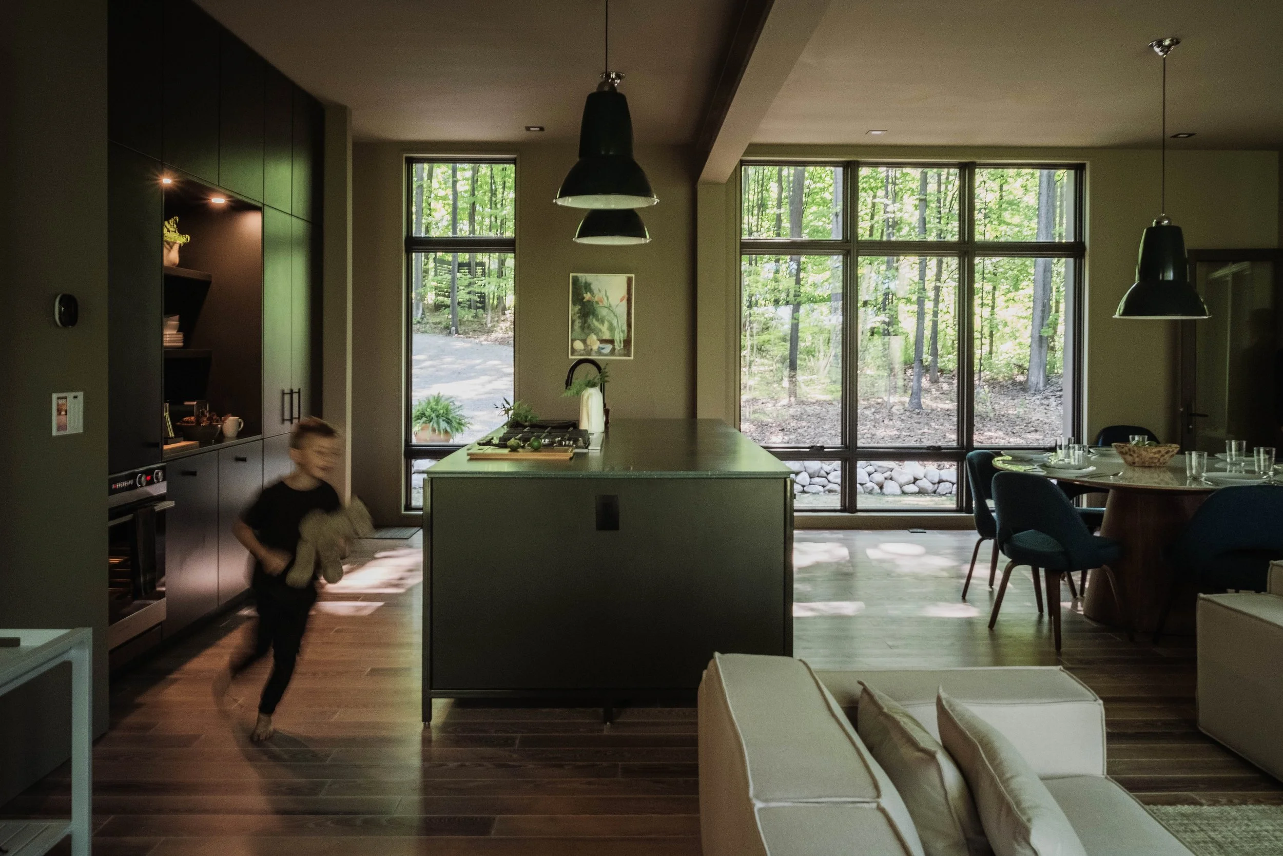 A modern kitchen with large windows showing a wooded outdoor scene, a kitchen island, and a dining area with chairs and table set for a meal. A blurred child runs through the space.