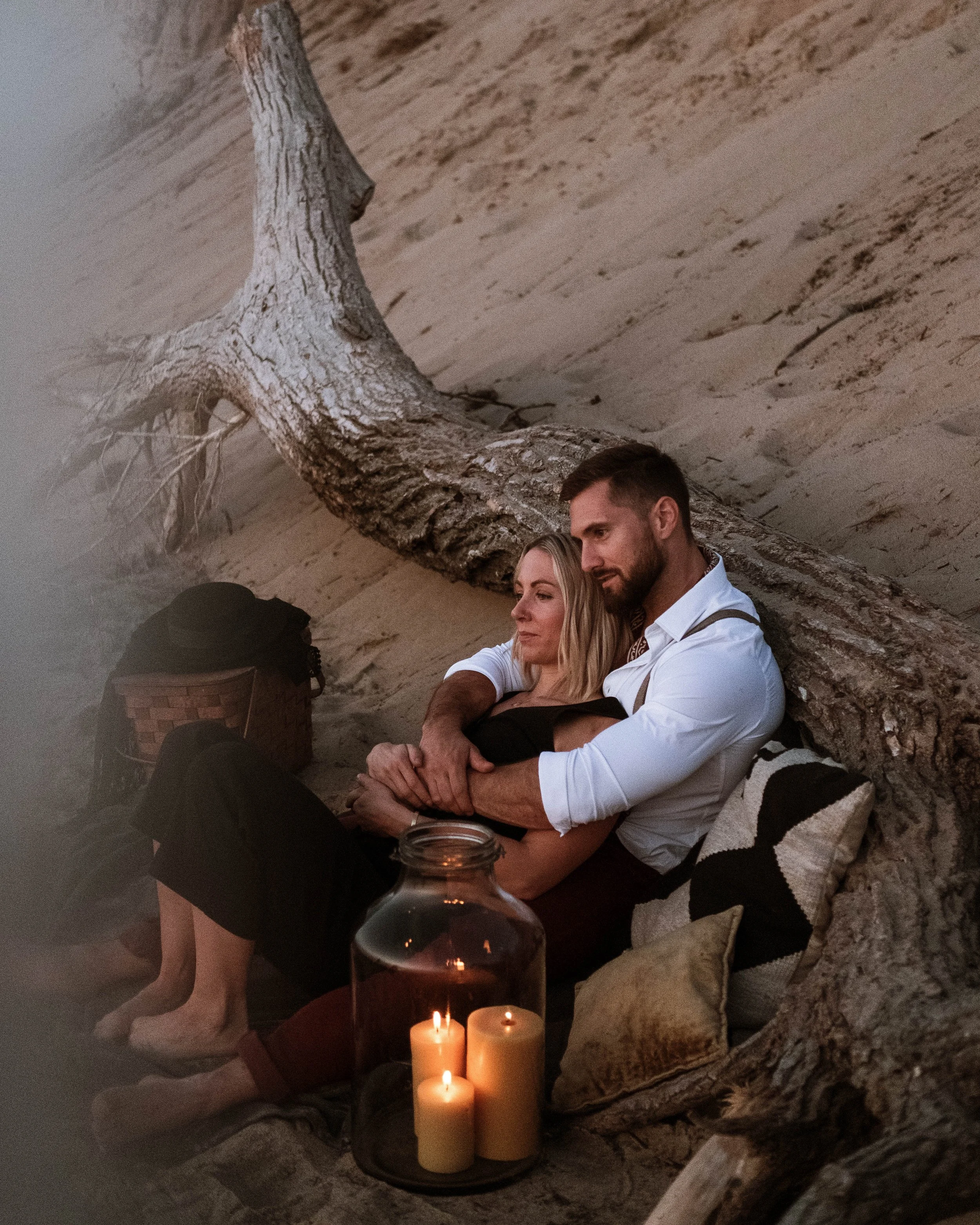 A couple sitting on the sand beneath a large fallen tree, with candles in a glass jar nearby, during sunset or evening.