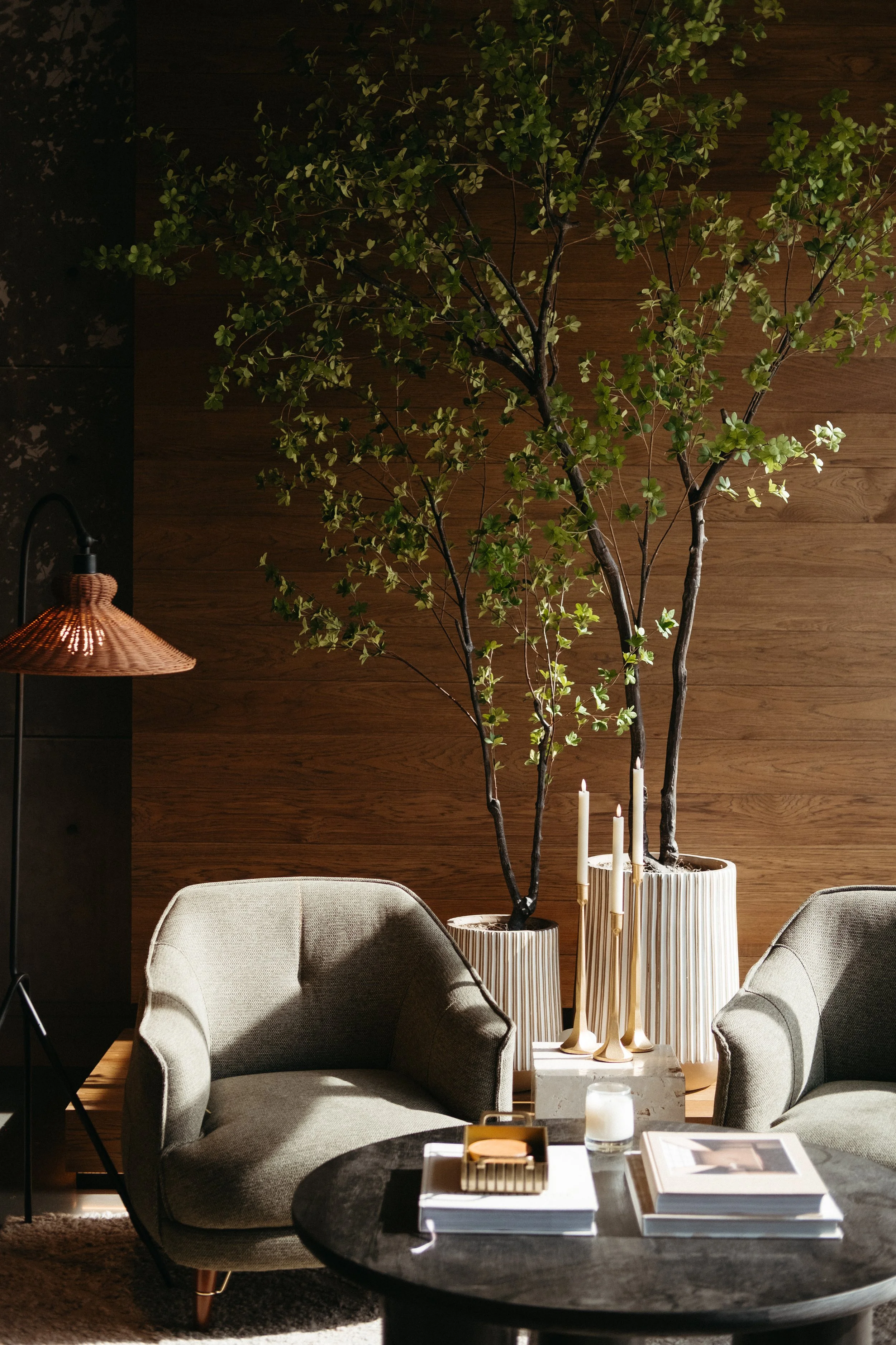 Living room with two beige armchairs, a dark round coffee table with books, candles, and a gold tray, and large potted plants with green leaves against a wooden wall, with a copper floor lamp on the left.
