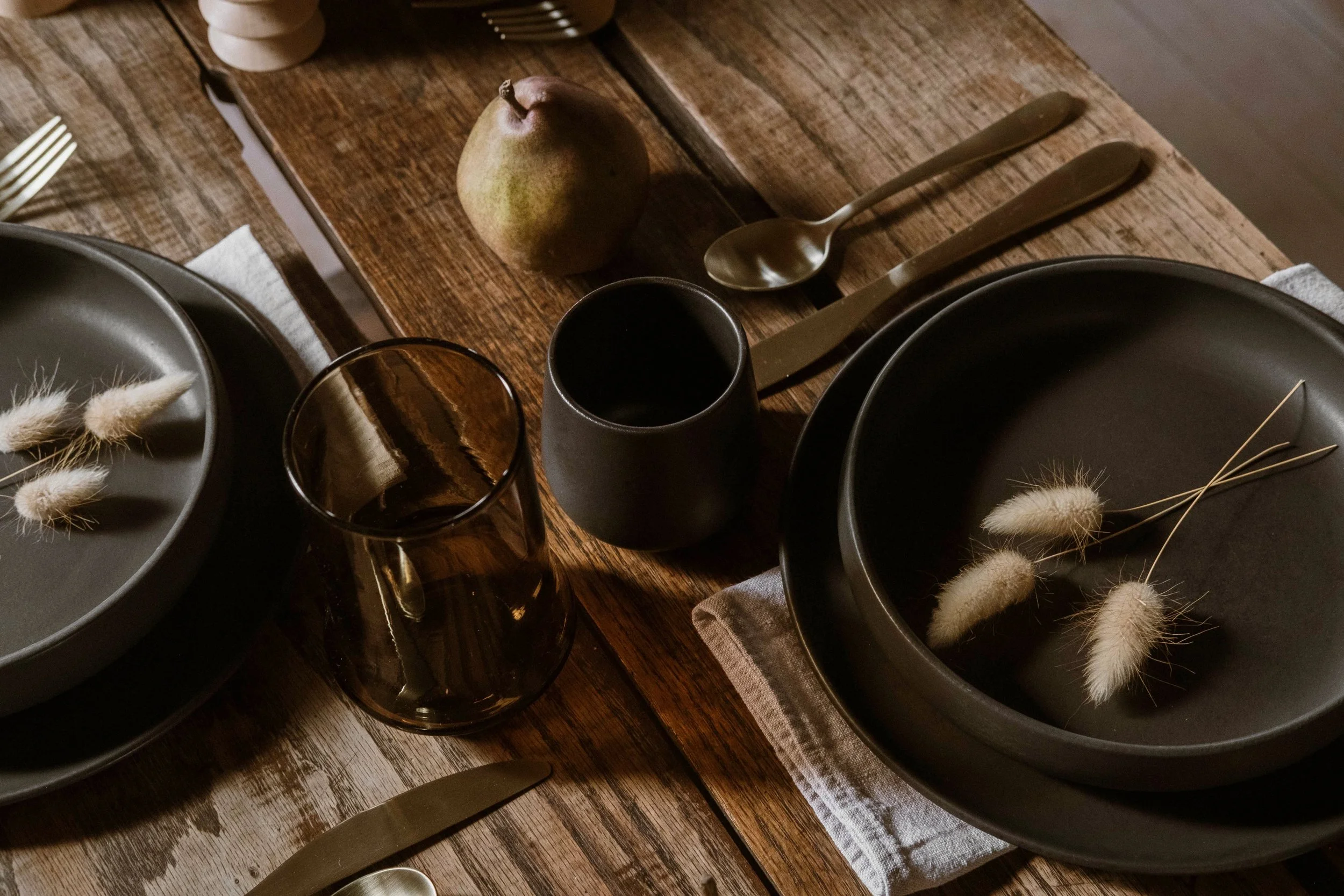 A rustic wooden table set with black dishes, a brown glass tumbler, a black cup, and gold-colored utensils. On the table are also a pear, some dried decorative wheat, and neutral-colored napkins.