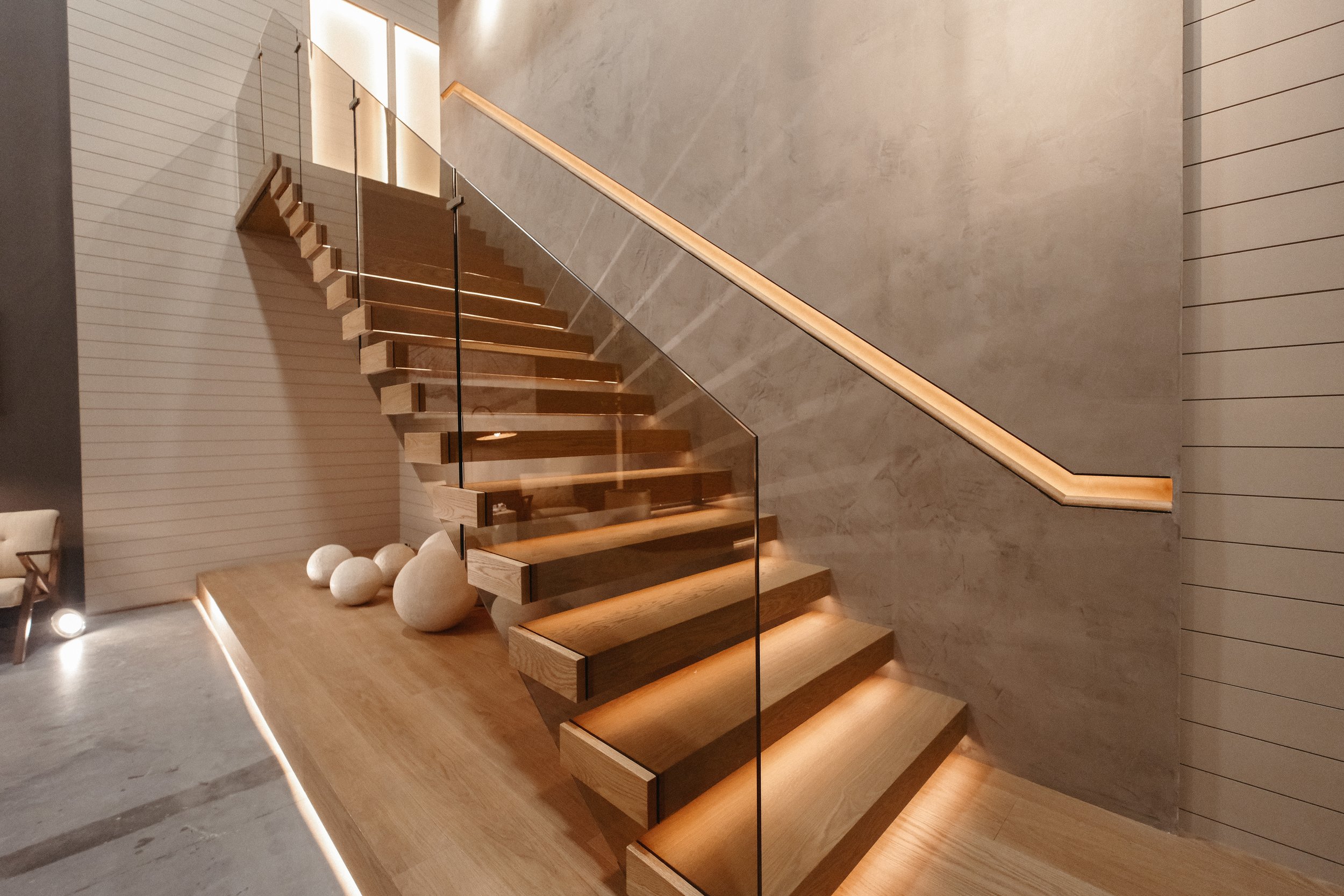 Modern wooden staircase with glass railing and built-in lighting, next to a wall with horizontal stripes and decorative white stones on a wooden platform.