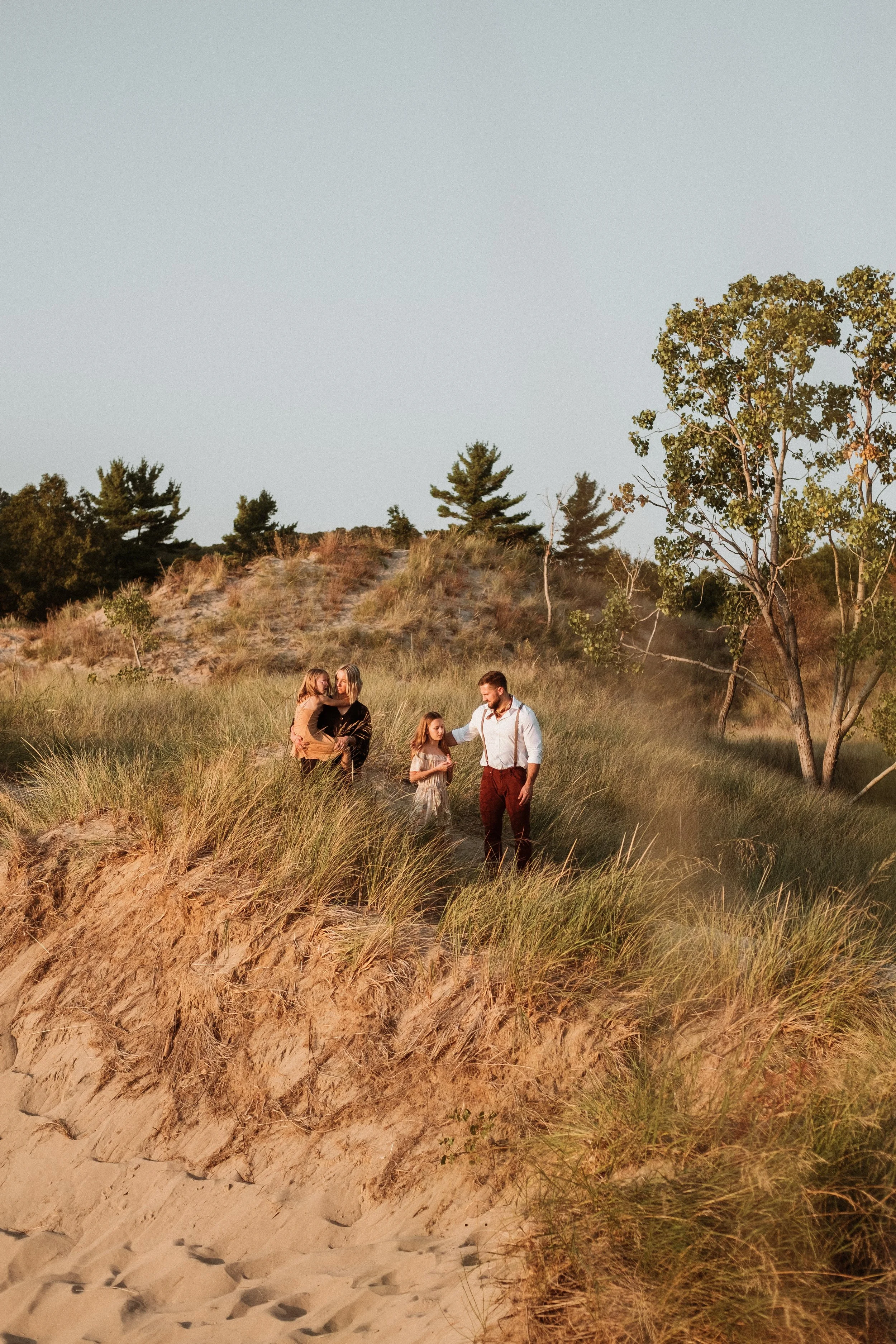 A family walking through grassy dunes near the beach at sunset, with trees in the background.