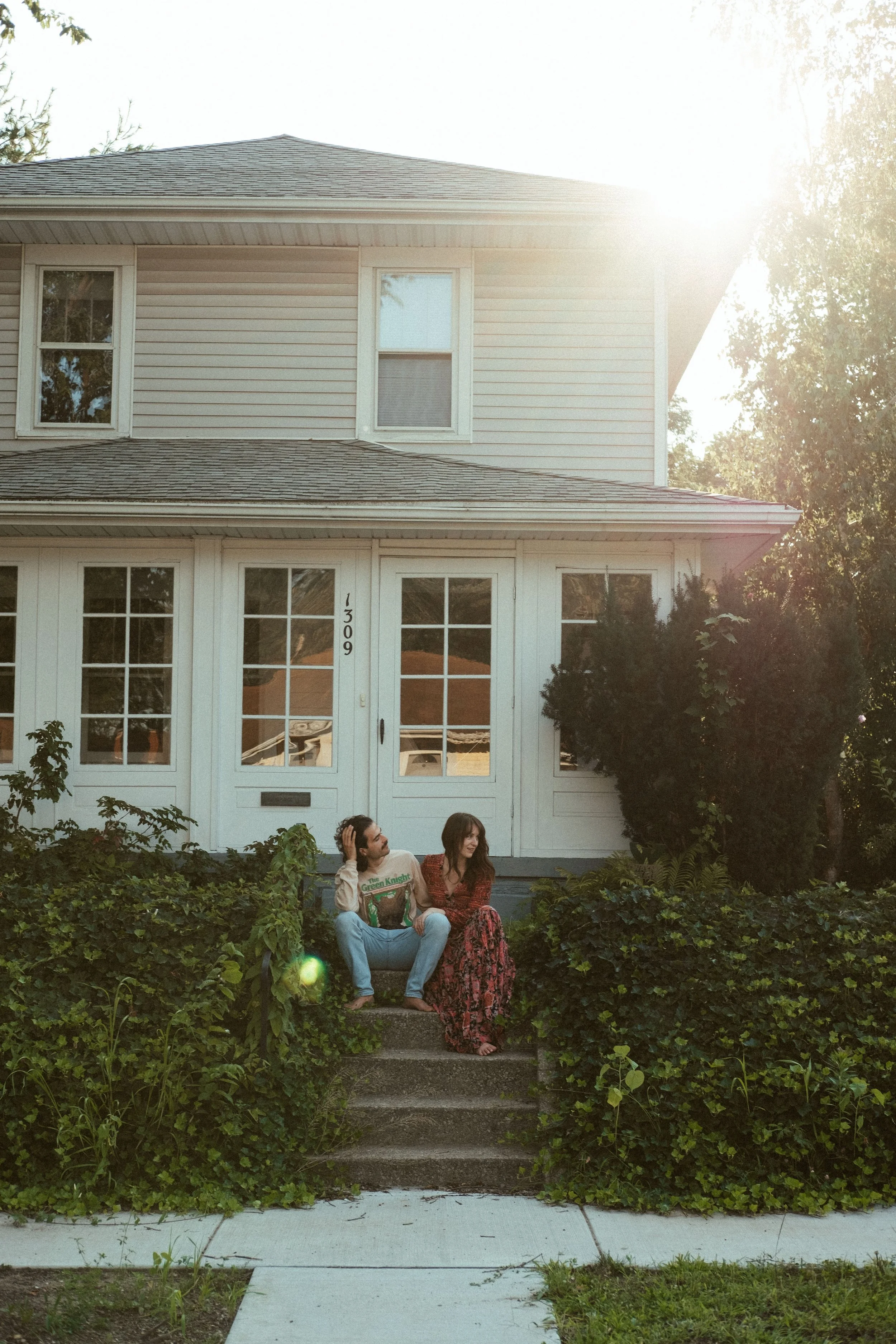 Two people sitting on stairs in front of a white house with a front porch, surrounded by greenery, during sunset.