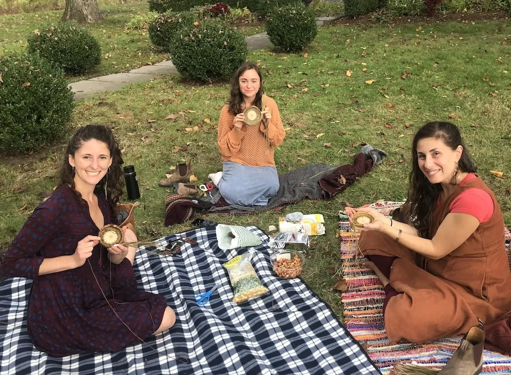 three women on a picnic blanket making pine needles baskets