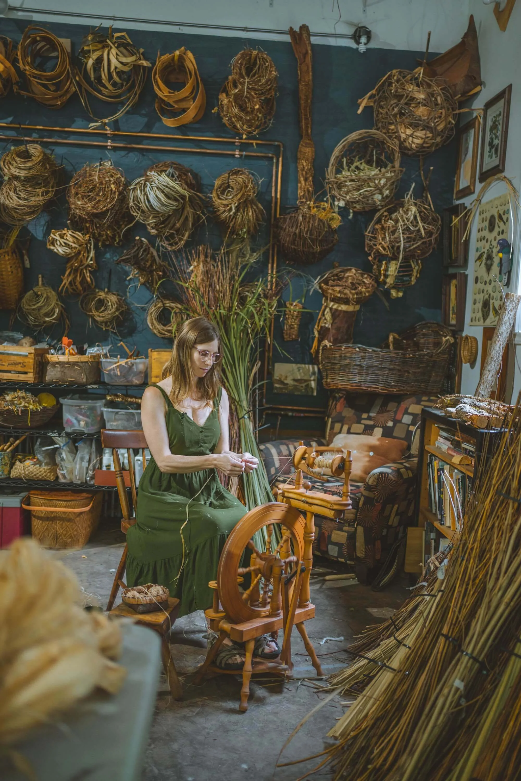 katie grove spinning basswood fiber on a spinning wheel in her studio with basketry materials like vines and bark on the studio wall