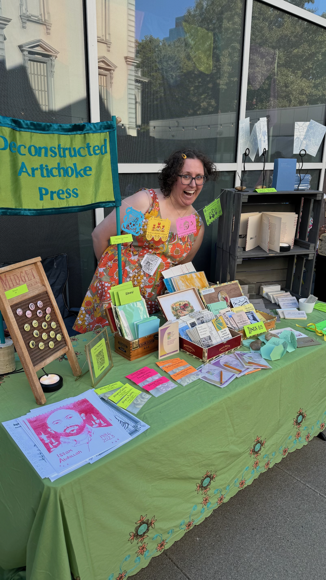 A woman at a booth with a sign that says 'Deconstructed Artichoke Press,' surrounded by books, pamphlets, and colorful paper products.