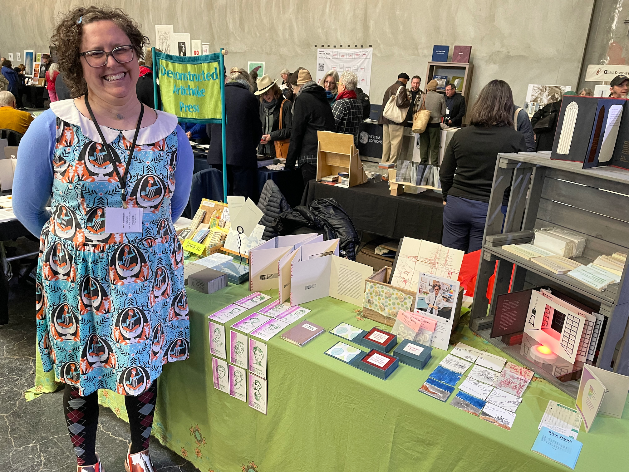 Woman smiling at a booth in a busy indoor market, with books, cards, and art prints displayed on the table, in front of a sign that reads "Deconstructed Artichoke Press".