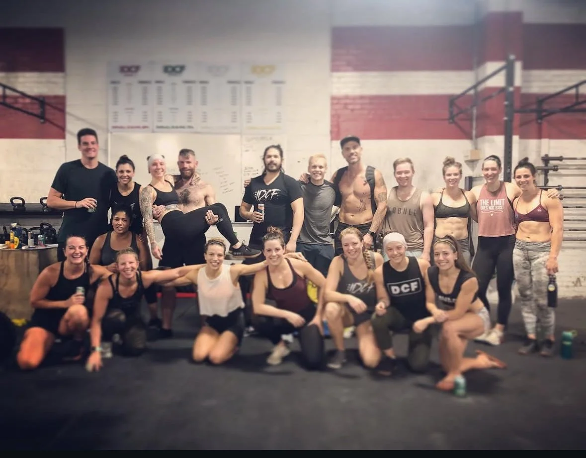 Group of people in workout clothes at a gym, some smiling and posing, with a whiteboard and fitness equipment in the background.