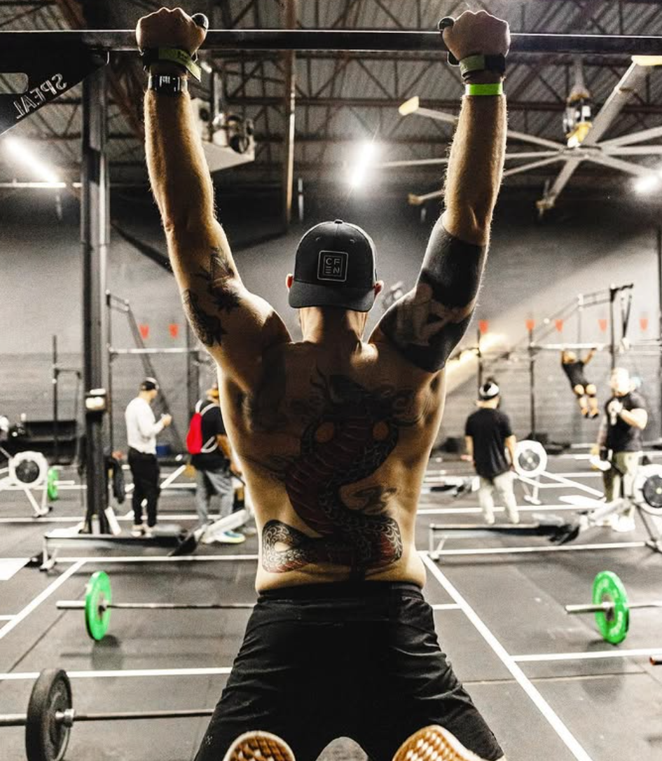 A shirtless man with tattoos on his arms and back, wearing a black cap, lifting a barbell overhead in a gym. Other people are in the background, also working out.
