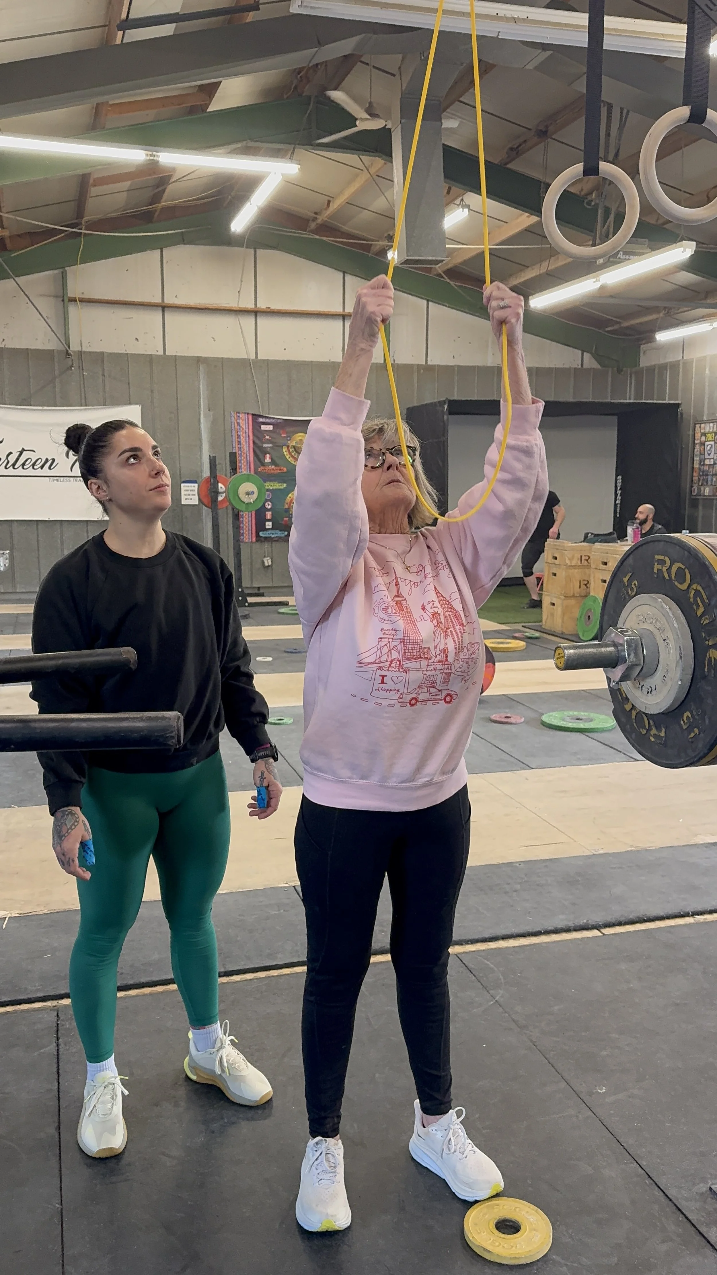 An elderly woman in a pink sweatshirt and black pants lifts a yellow jump rope overhead in a gym, with a younger woman standing nearby holding a barbell and watching her. The gym has weightlifting equipment, hanging gymnastic rings, and weight plates on the floor.