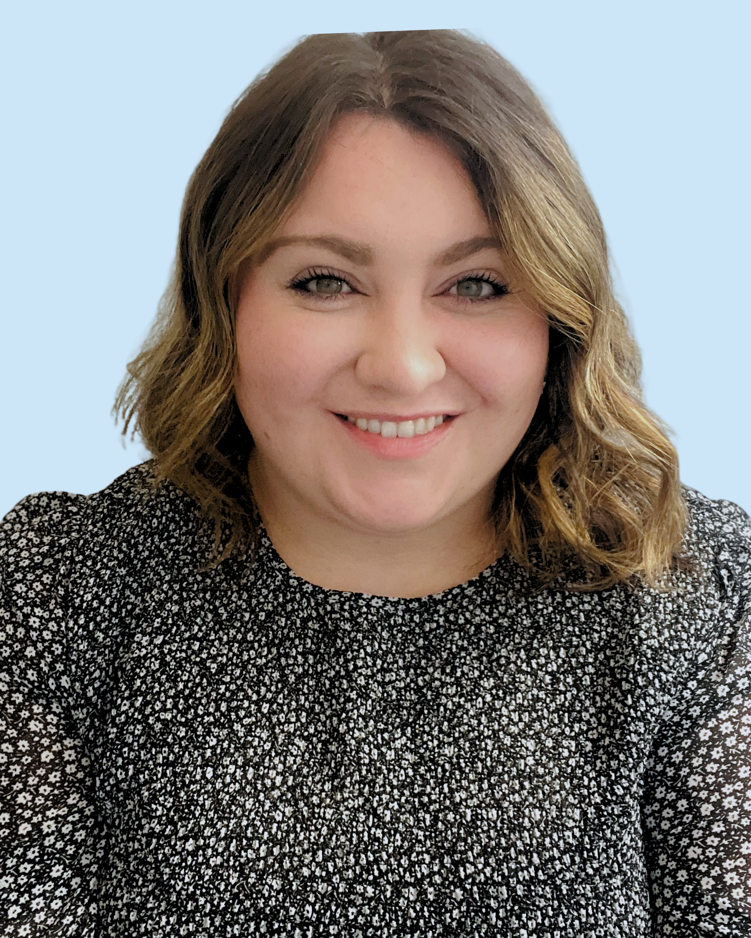 A woman with shoulder-length wavy brown hair, light skin, and blue eyes smiling at the camera, wearing a black and white floral patterned top, against a light blue background.