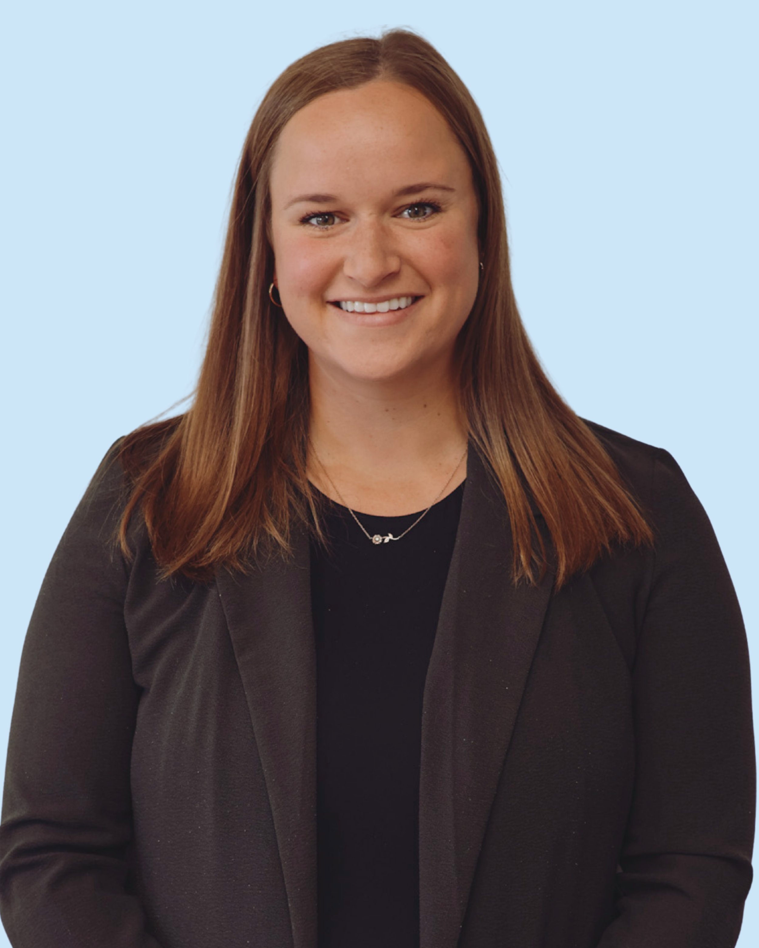 A woman with straight, shoulder-length auburn hair, smiling, wearing a black blazer, black top, and a necklace with a small charm.