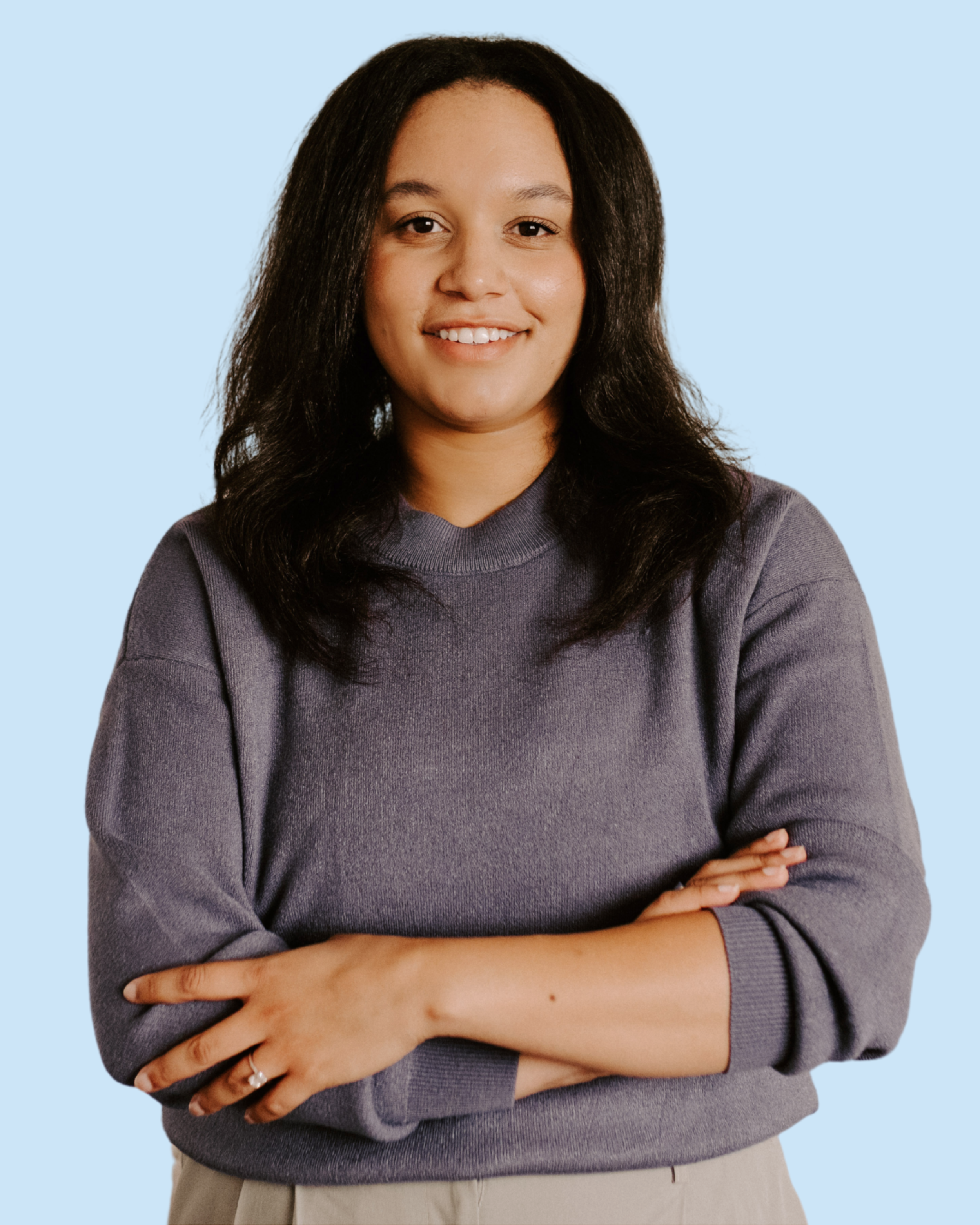 A woman with medium skin tone, black hair, wearing a gray sweatshirt and beige pants, standing with arms crossed in front of a light blue background.