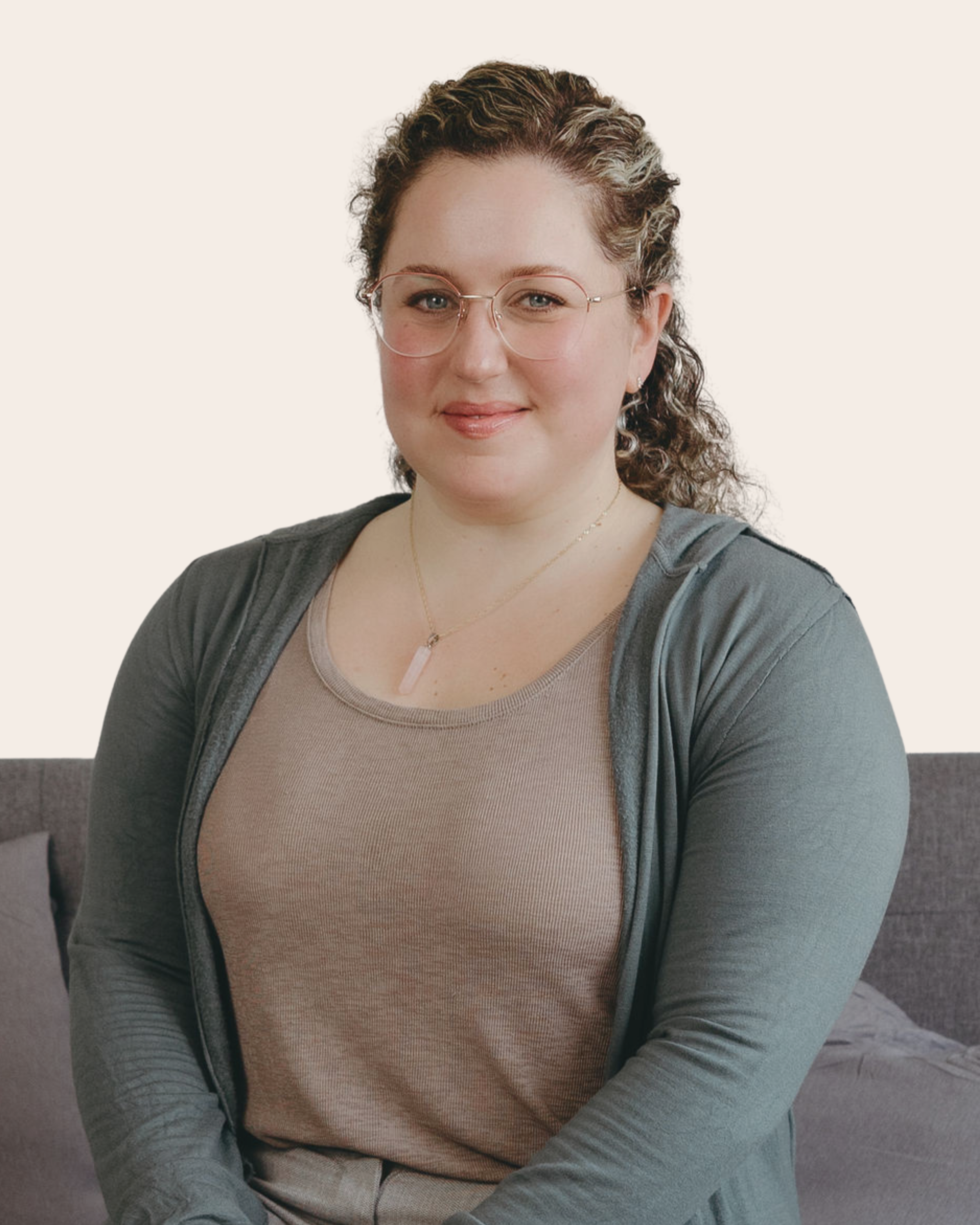 A woman with curly hair, glasses, and a warm smile sitting on a gray couch in front of a plain light background.