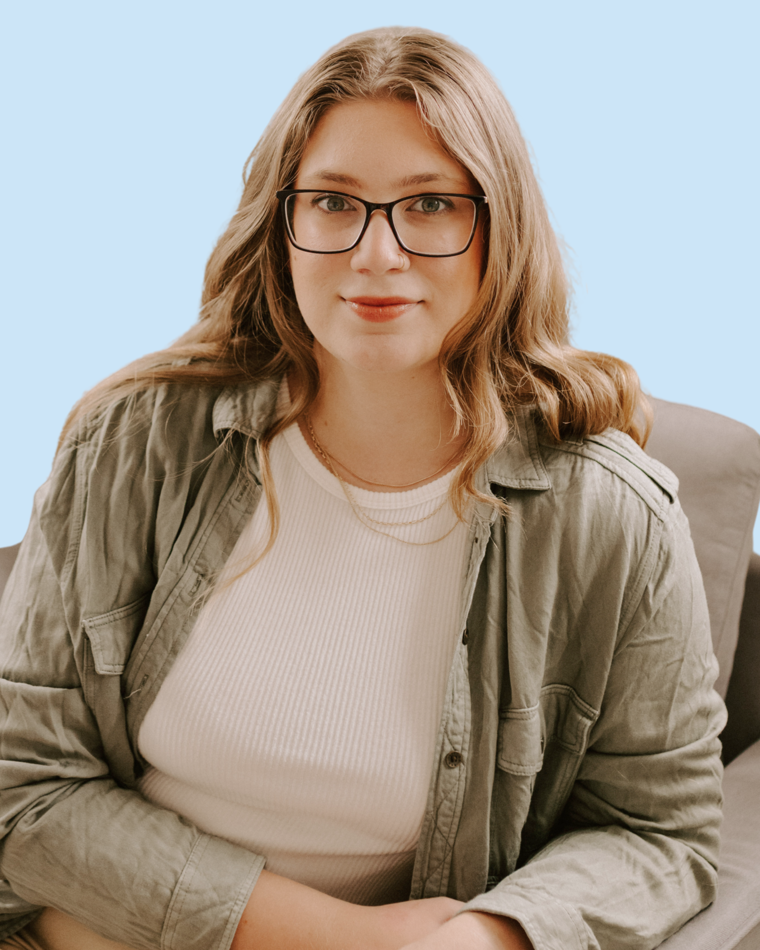 A young woman with wavy blonde hair, glasses, and a confident smile sitting on a beige chair against a light blue background.