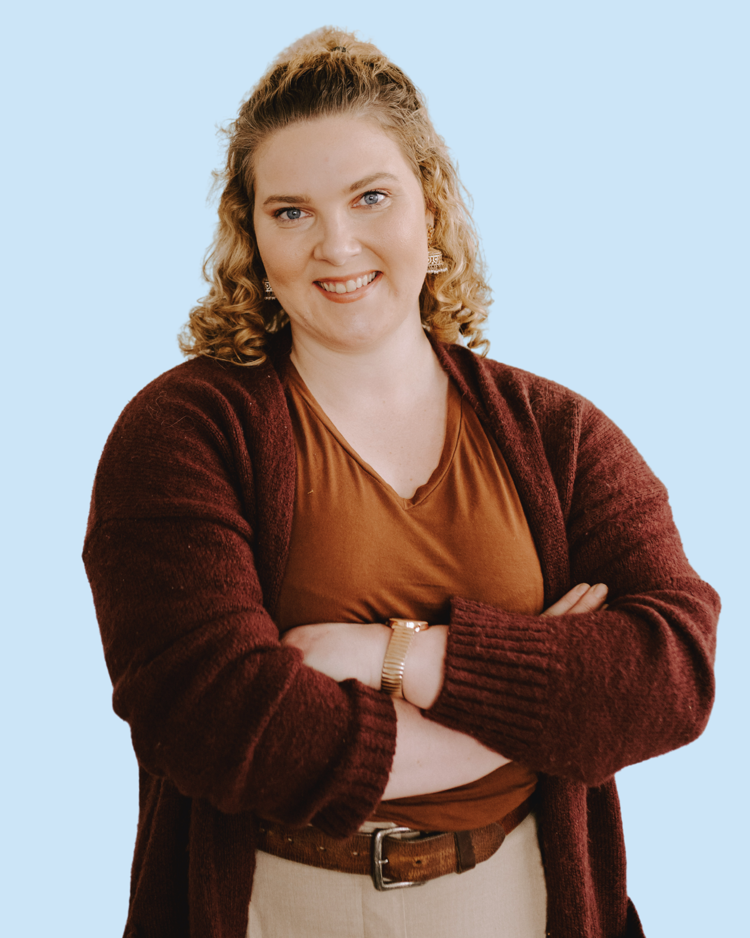 A woman with curly blonde hair, wearing a brown shirt and a maroon cardigan, crossing her arms and smiling against a light blue background.